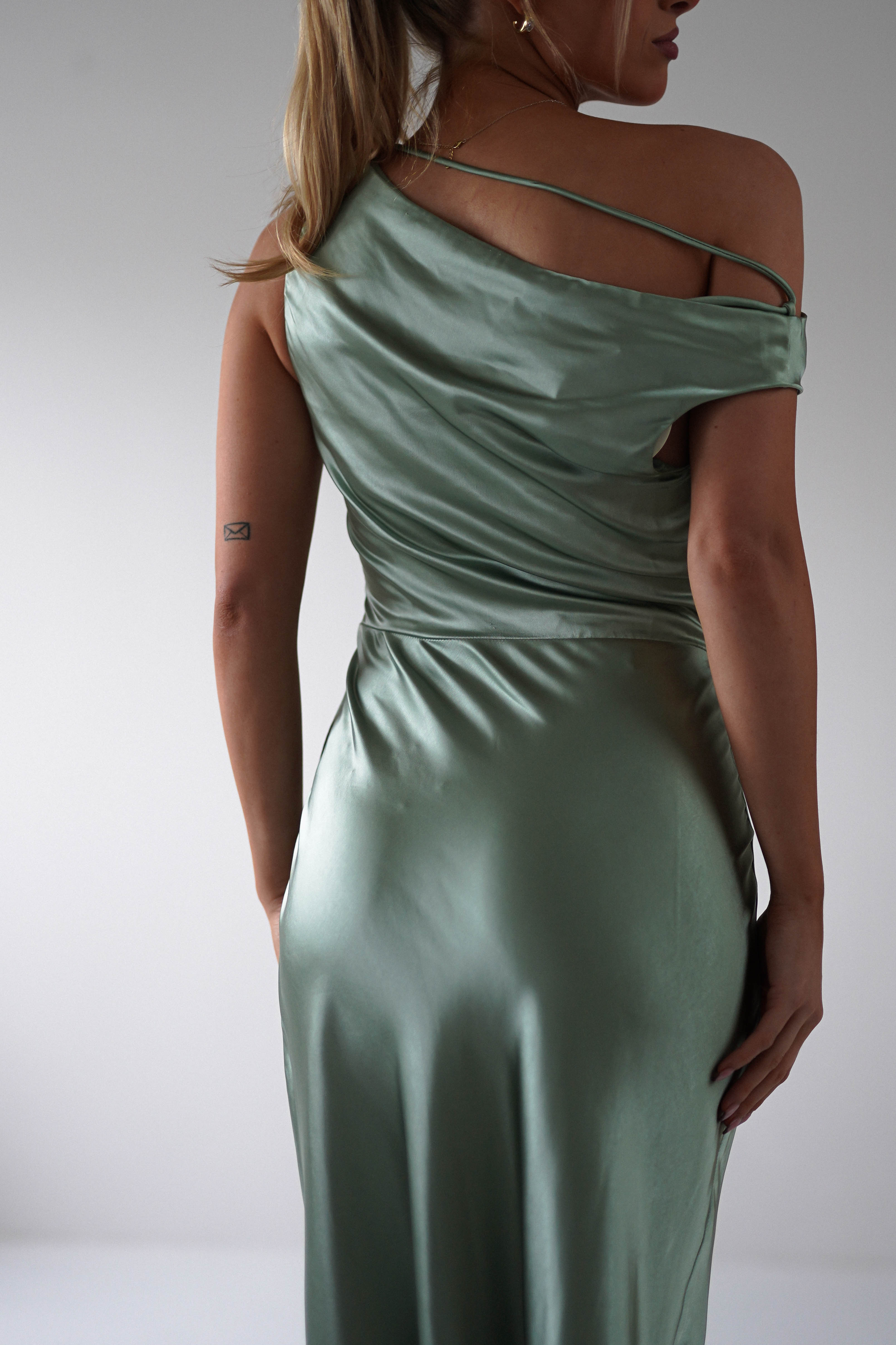 A person wearing a sleek, off-shoulder, sage green satin dress with a small tattoo on their arm stands turned slightly away, their long hair tied back. Perfect for bridesmaids or any wedding guest, the dress drapes elegantly and reflects light softly.