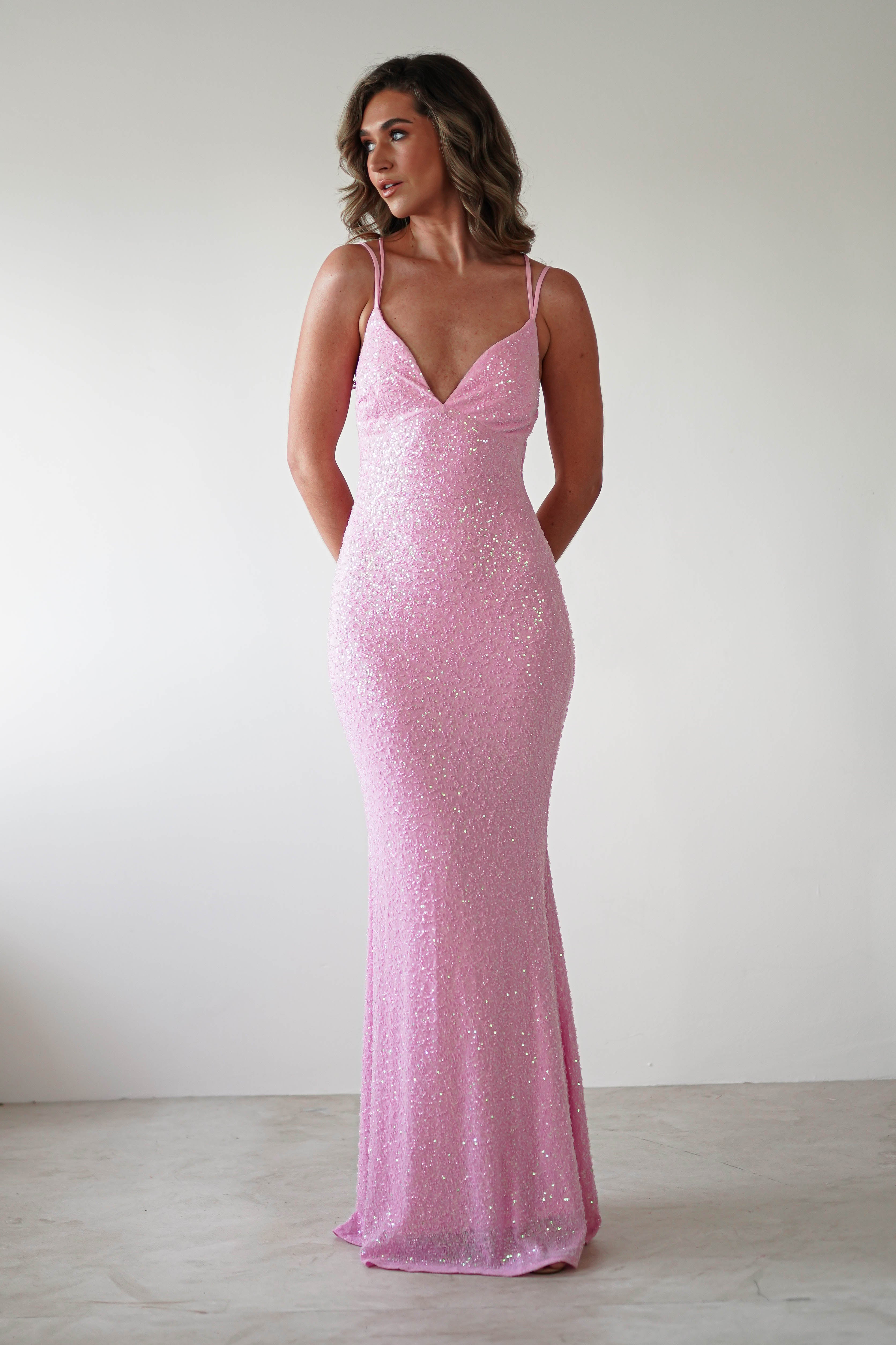 A person in a sparkly pink sequin gown with a graceful V-neckline stands against a plain white background. The dress is form-fitting, ideal for special occasions, and boasts an elegant silhouette. The person has long wavy hair and is looking slightly to the side.