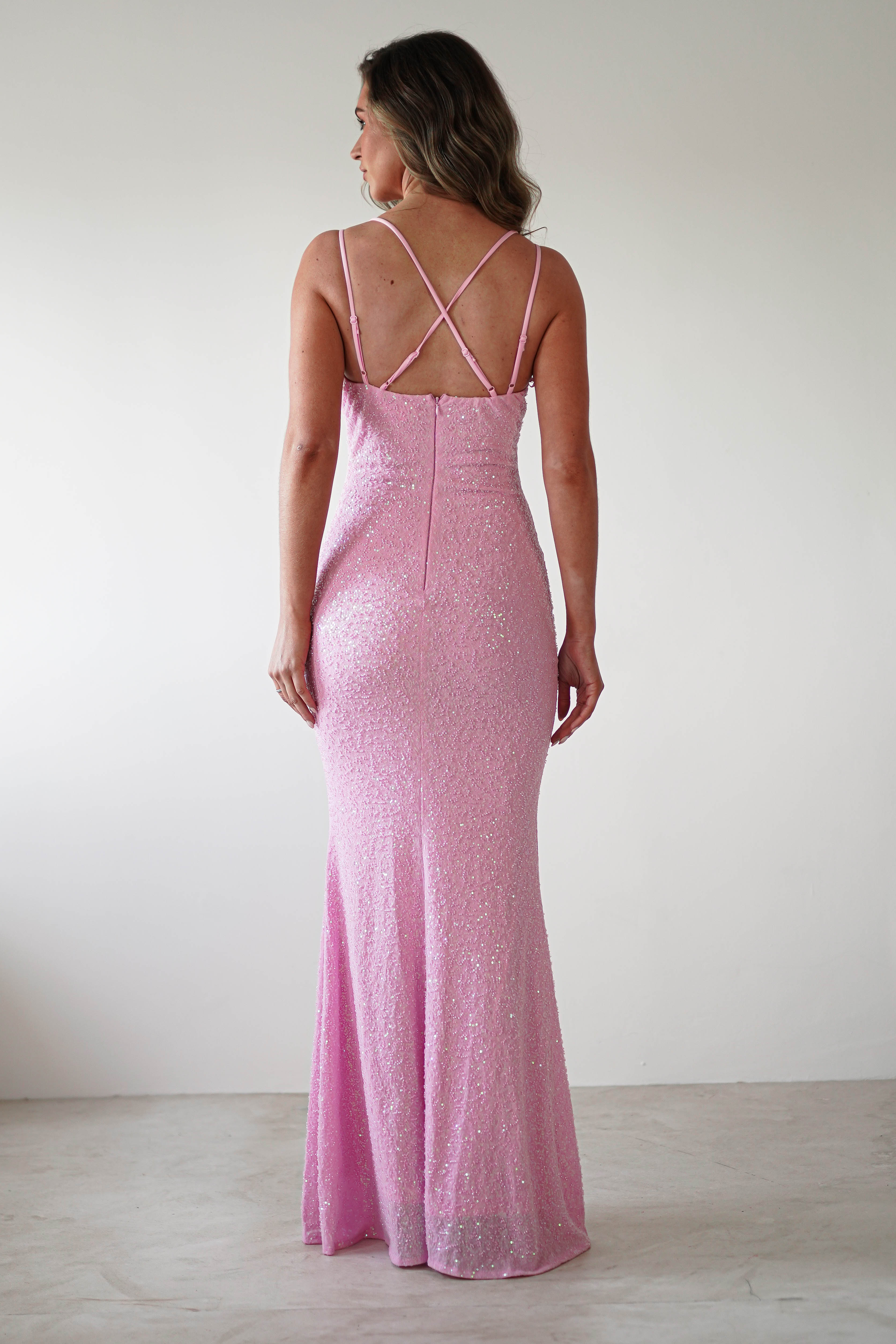 A woman stands facing a white wall, wearing a glittery pink sequin gown. The evening dress features thin straps, a V-neckline, and a fitted silhouette perfect for special occasions. Her hair is styled in loose waves, with the gowns back showcasing a zipper and crisscross straps.