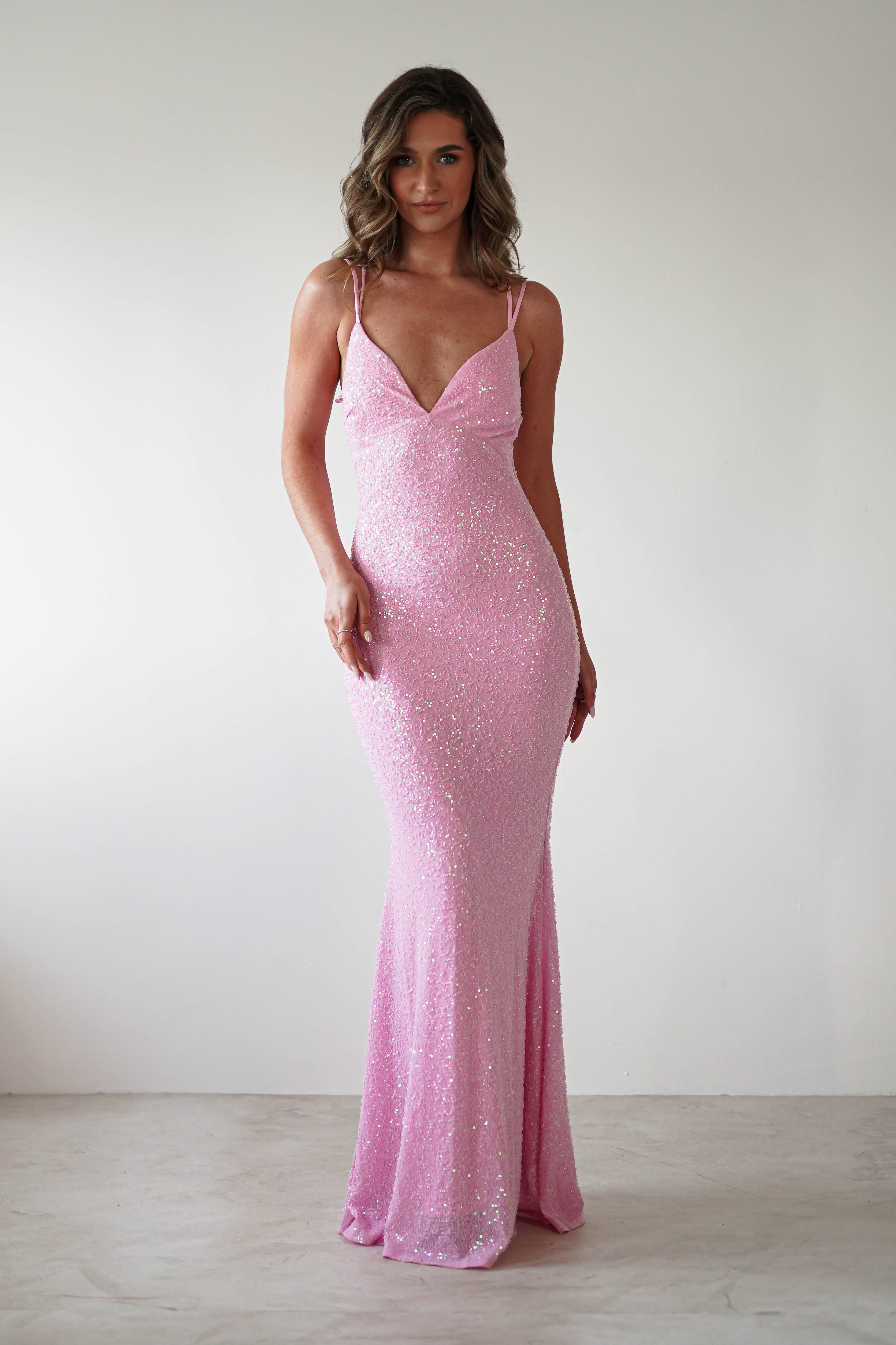 A person poses in a fitted sequin gown with thin straps and a deep V-neckline, perfect for special occasions. The sparkling pink dress gracefully flows to the floor as they stand against a plain white background.