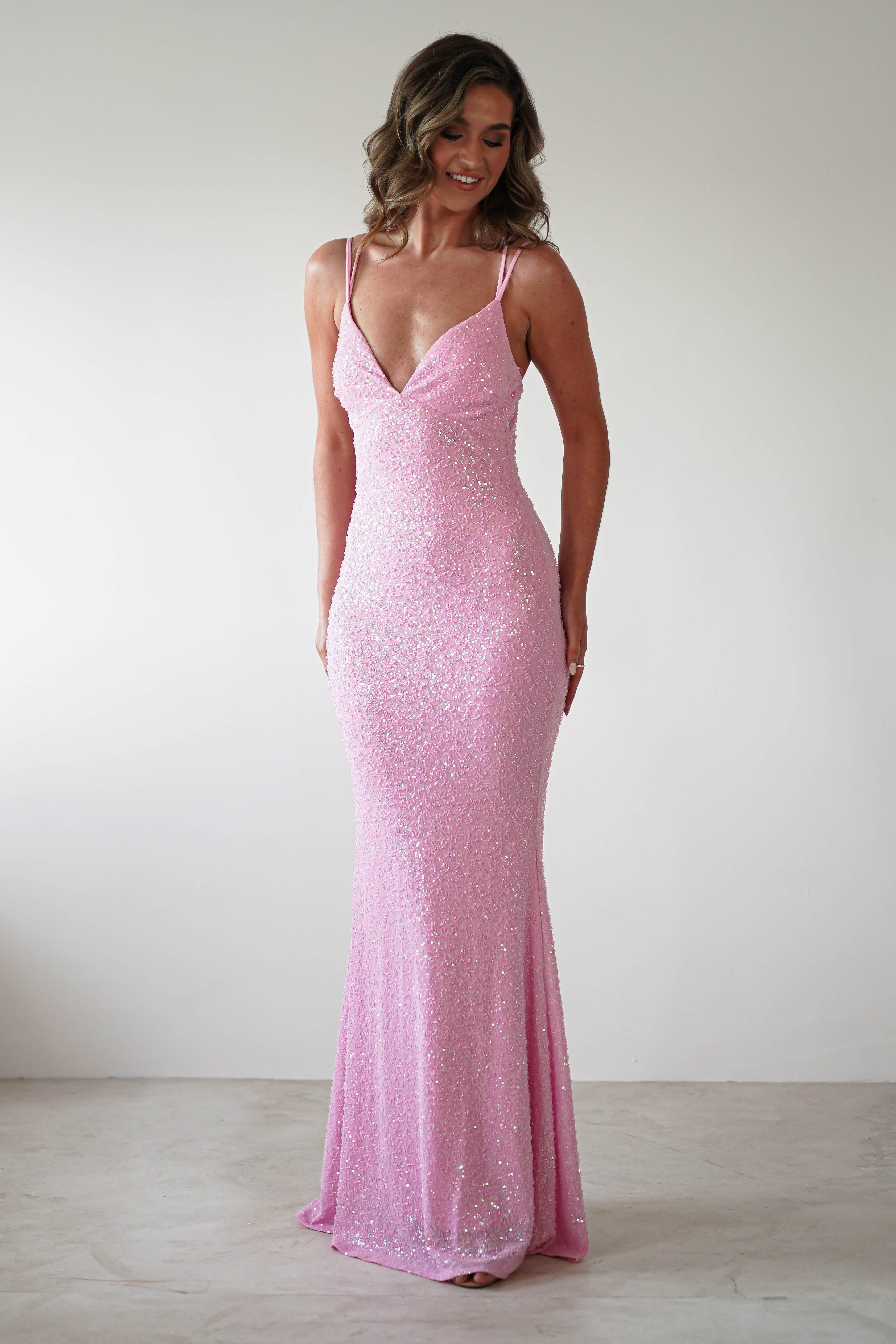 A person wearing a shimmering sequin gown stands against a plain background. The sparkling pink dress, perfect for special occasions, features thin straps and a V-neckline, accentuating their figure. They have wavy hair and a gentle smile, looking downwards.