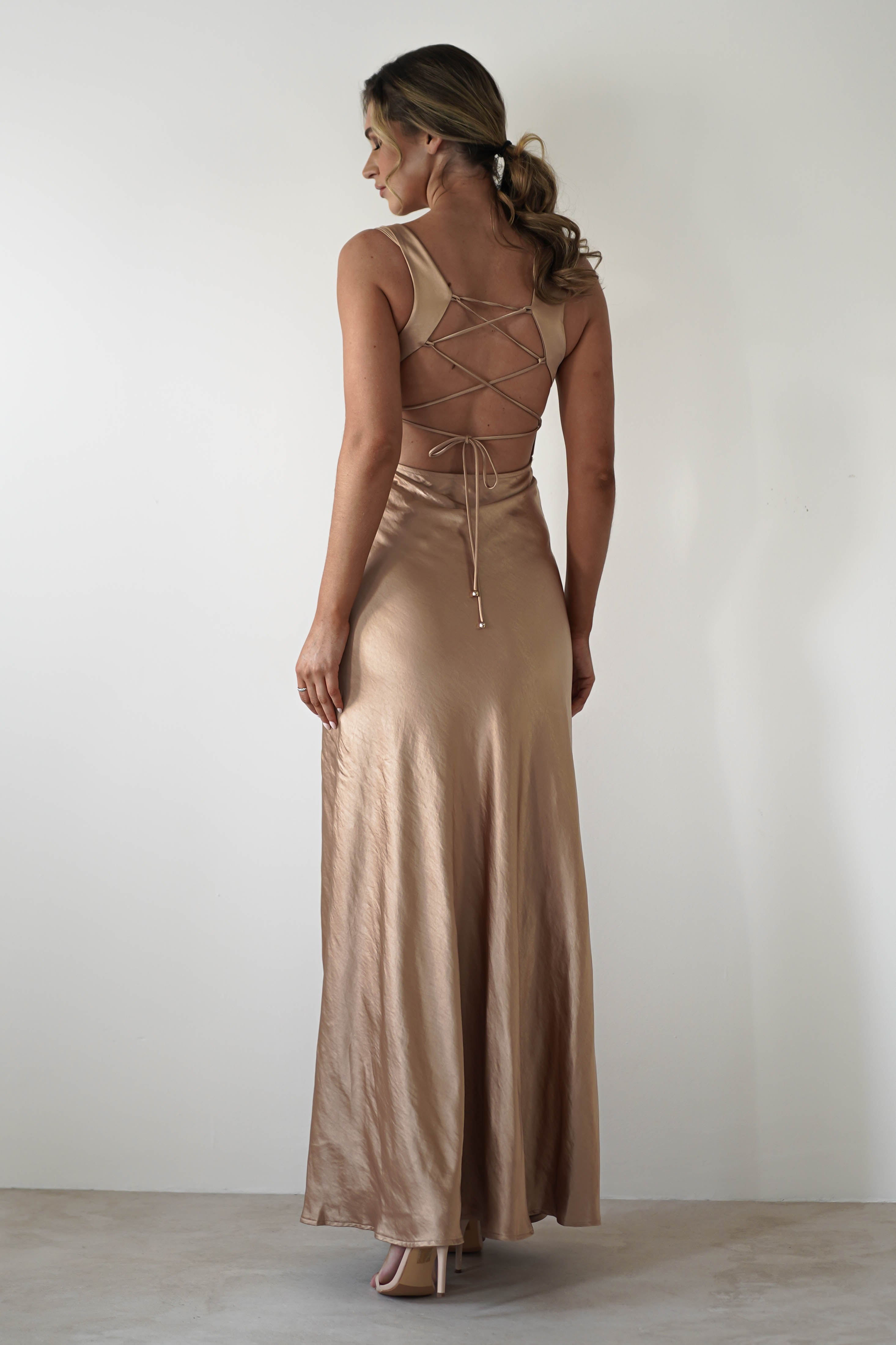 A woman stands against a plain white wall, wearing a gold satin gown with a cowl neckline and an open back featuring crisscross lace-up detailing. She has wavy hair pulled back and is looking to the side.