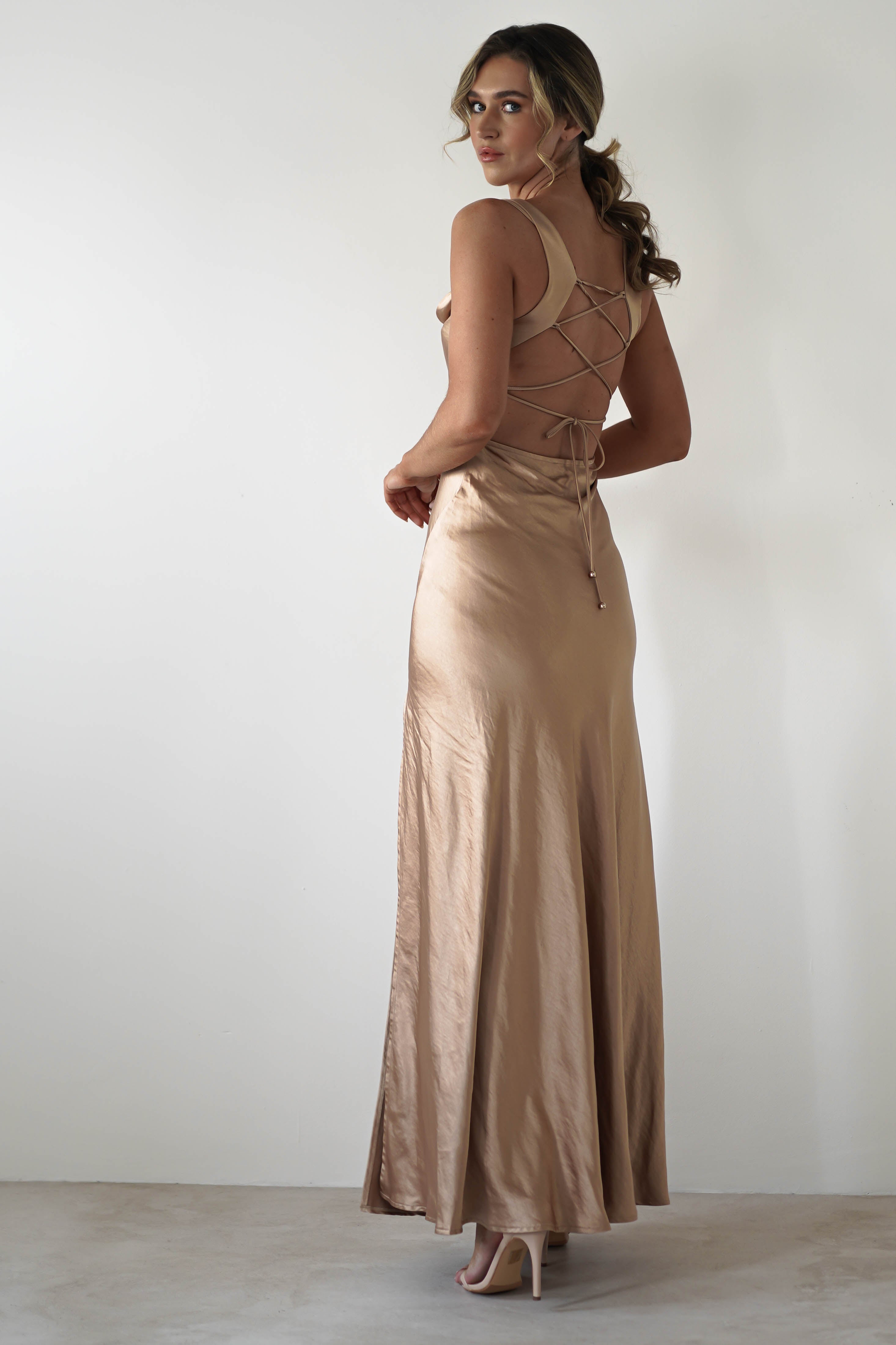 A woman wearing a long, silky satin gown with a cowl neckline and lace-up open back stands in front of a plain white wall, looking over her shoulder. She has wavy hair styled in a low ponytail and is wearing high heels.