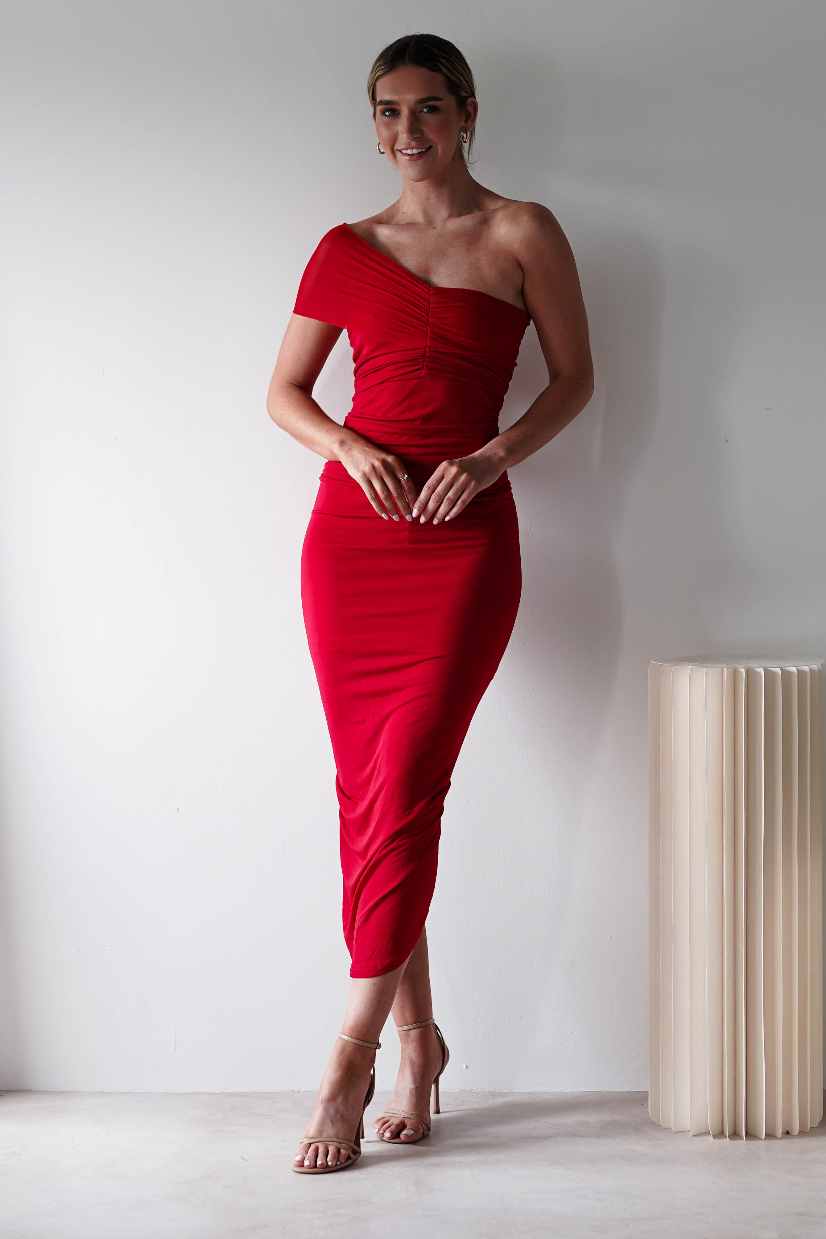 A woman stands indoors against a plain white wall, wearing a ruched bodycon, off-the-shoulder red dress and nude heels—an elegant look thats perfect for a wedding guest. Her hair is pulled back, and a sculptural object rests beside her.
