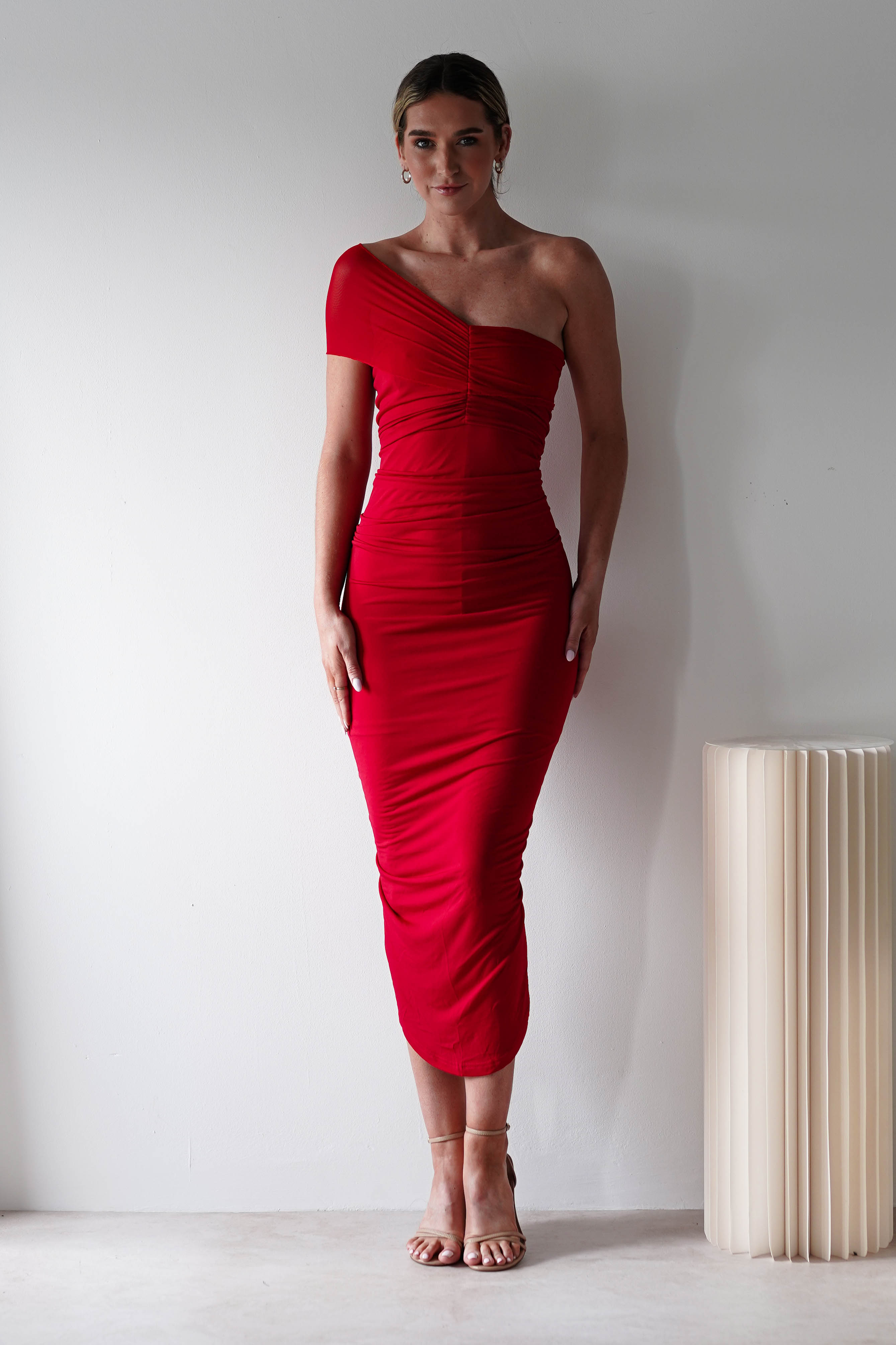 A woman stands against a white wall wearing a fitted, ruched bodycon red one-shoulder dress and nude heels. She is facing forward with her hands by her sides. A cream-colored sculptural table is on her left—perfect for a chic wedding guest look.