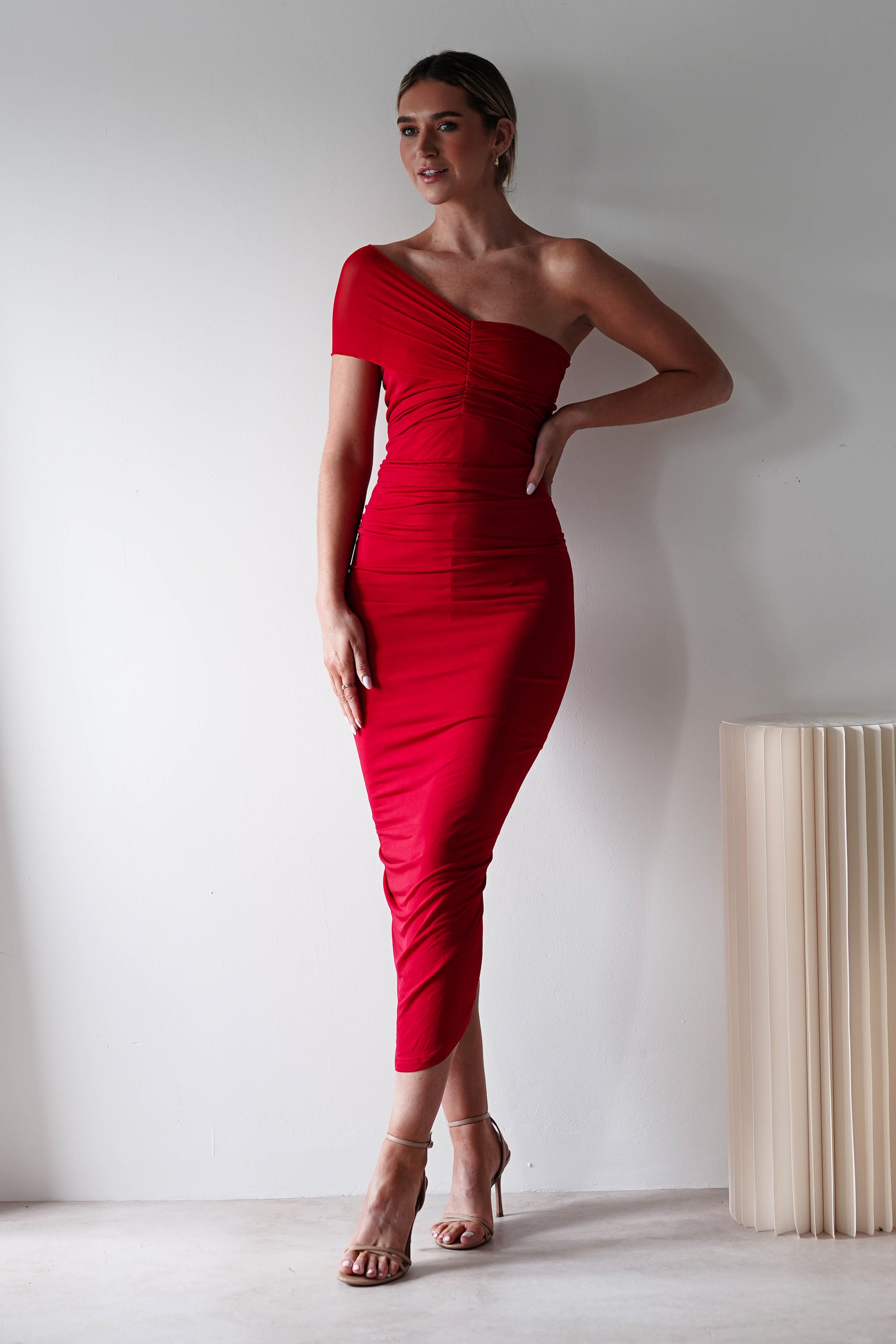 A woman stands against a white wall wearing a ruched bodycon, off-the-shoulder red dress and strappy heels, her hand on her hip. A cream-colored, modern floor lamp is visible beside her—perfect for a stylish wedding guest look.
