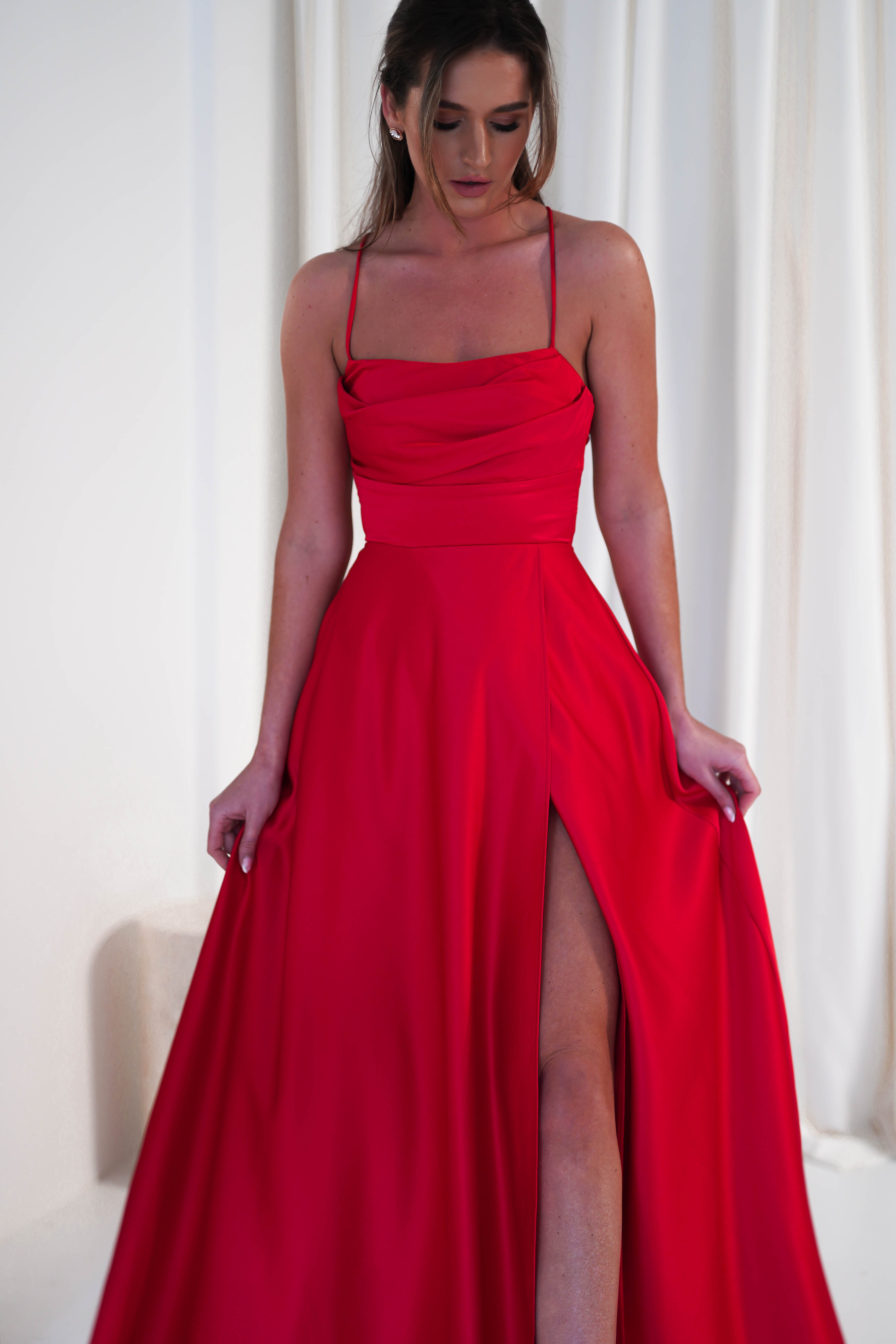 A person wearing a long, elegant satin gown with thin straps and a cowl neckline stands gracefully against a light curtain background, the thigh-high slit adding a touch of allure.