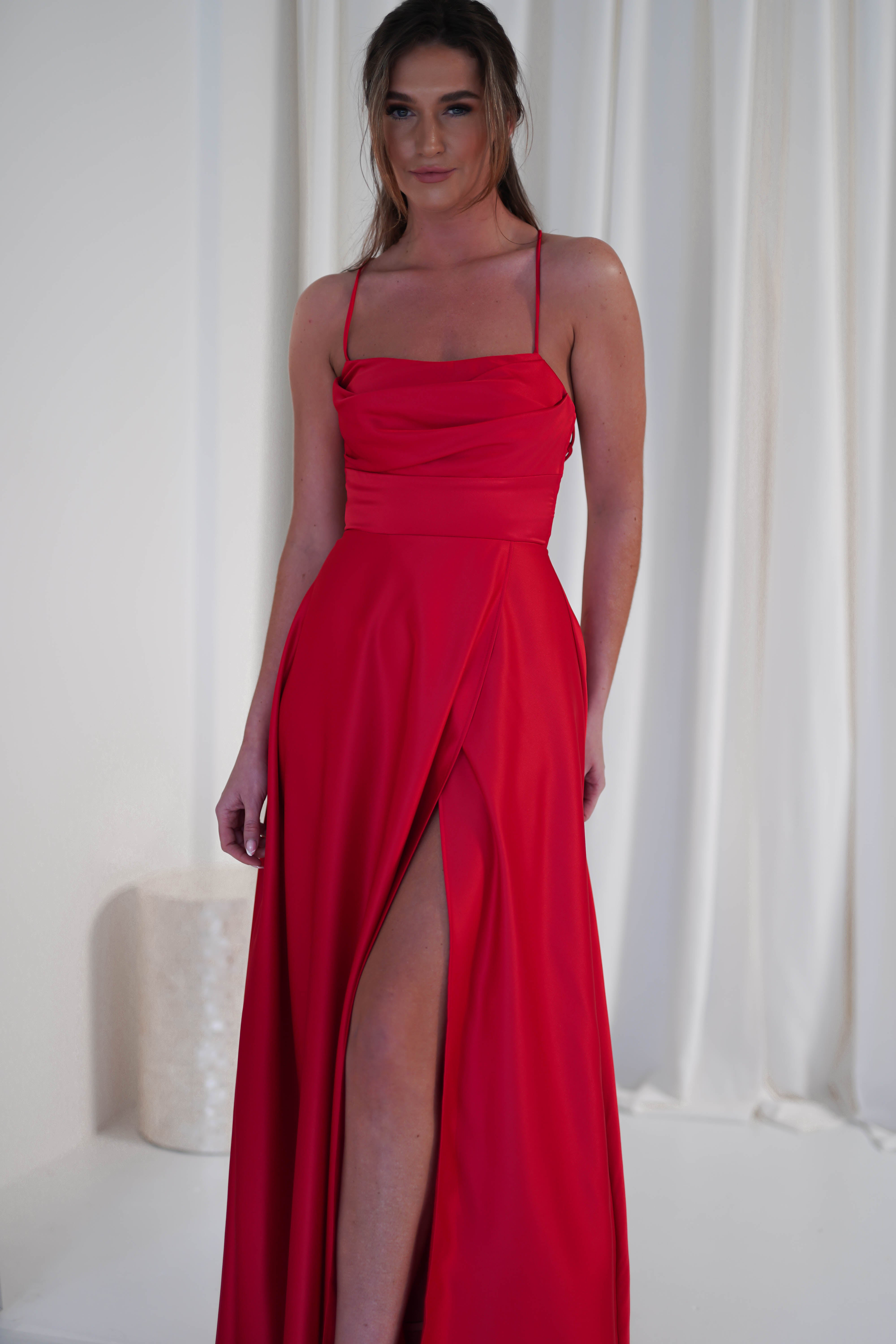 A woman in a red satin gown with a cowl neckline and a thigh-high slit stands in front of a white curtain. She is smiling and has her hair styled back.