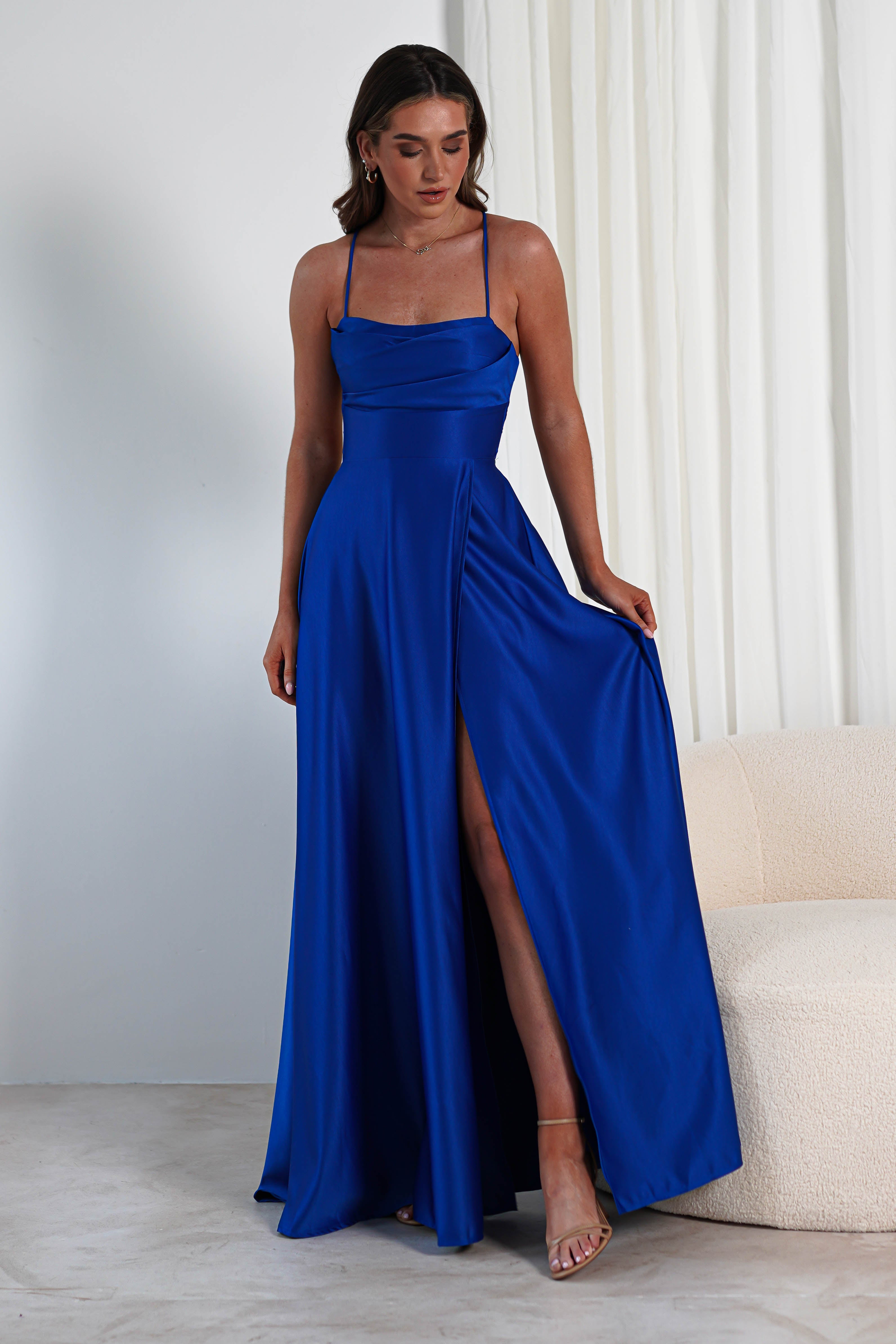 A person wearing a long, elegant satin gown with spaghetti straps and a cowl neckline stands in a minimalistic room. The dress features a thigh-high slit, revealing one leg. The individual poses beside a light-colored sofa with white curtains in the background.