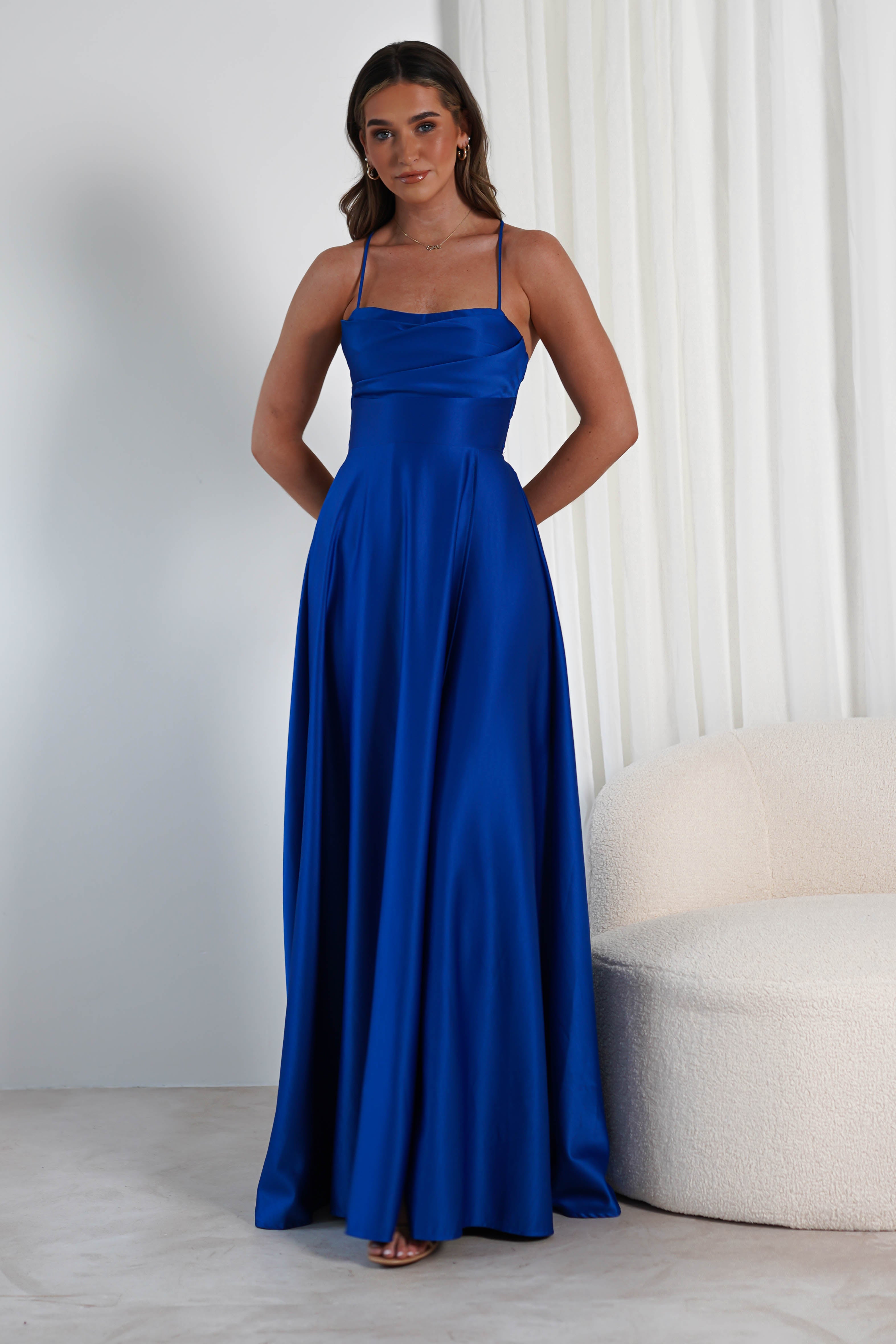 A woman stands gracefully in a blue satin gown with spaghetti straps and a fitted bodice. The dress features a subtle cowl neckline, complementing the flowing skirt where her hands rest in the pockets. She is set against a modern backdrop with a light-colored armchair and curtains nearby.