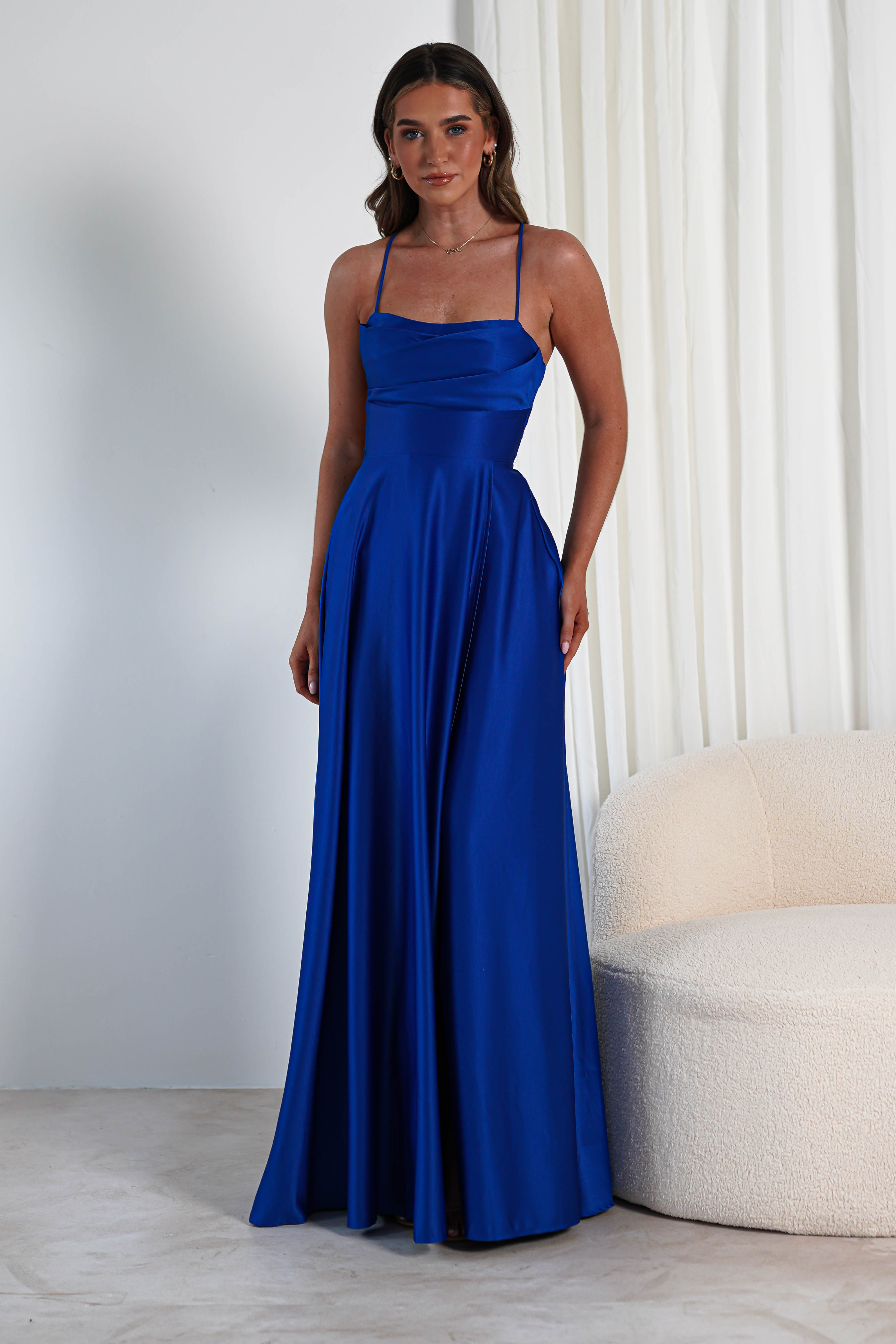 A woman stands in a blue, floor-length satin gown with a cowl neckline and spaghetti straps. The fabric is smooth and slightly glossy. She is indoors, beside a white curtain and cream-colored sofa, with the gowns thigh-high slit subtly revealing her leg.