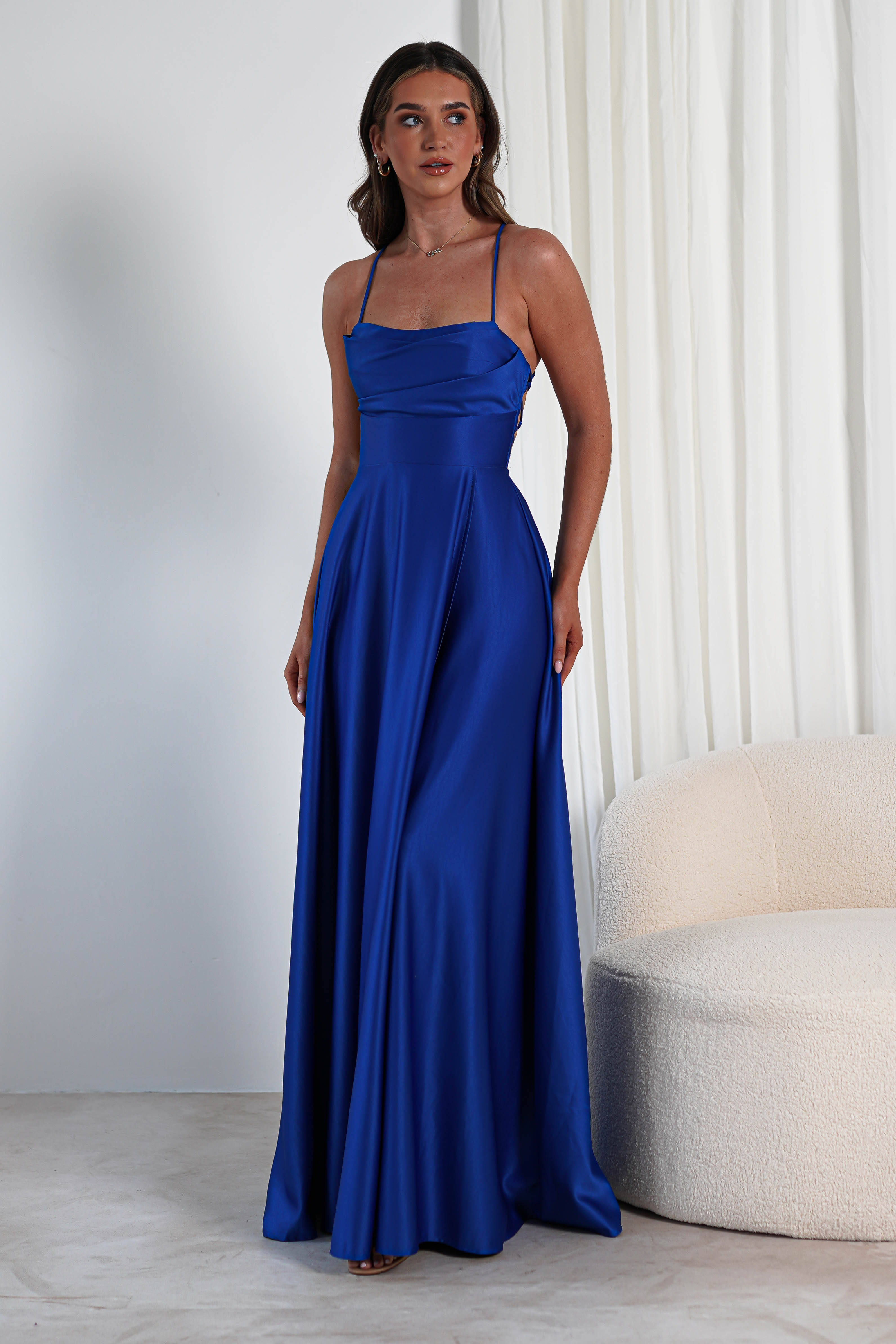 A woman in a stunning blue satin gown stands beside a cream-colored, textured sofa. The dress features thin straps, a cowl neckline, and a thigh-high slit in the flowing skirt. She poses in front of soft, white curtains, creating an elegant and serene atmosphere.