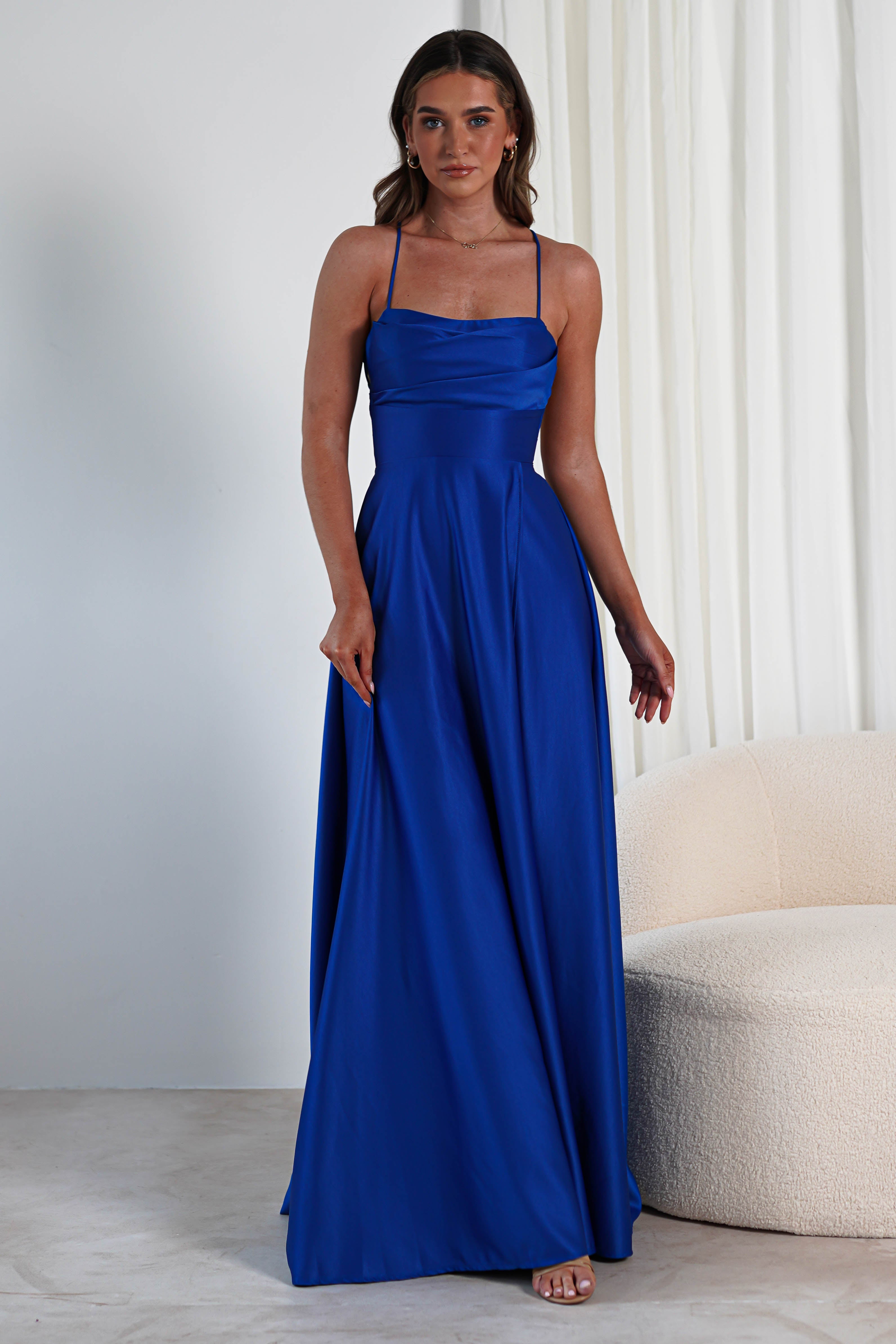 A woman stands in a room wearing a long, elegant blue satin gown with thin straps and a cowl neckline. Her hands gently touch the fabric as a thigh-high slit subtly reveals her silhouette. The background features white curtains and a beige, textured chair.