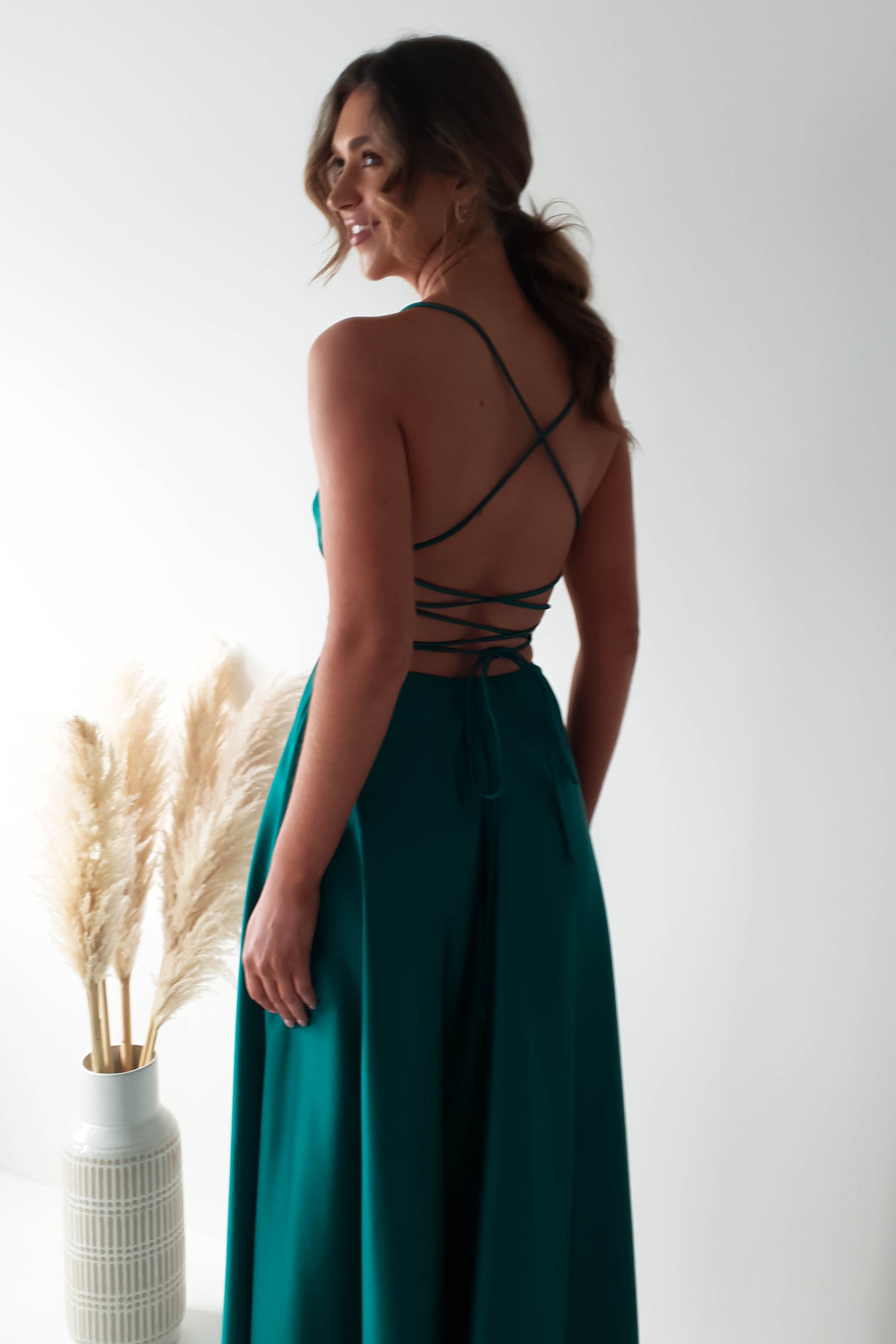 A woman in a teal dress with a cowl neckline and strappy back design stands with her back to the camera. Her long hair is tied back, revealing the dresss elegant silhouette. Nearby, a vase with tall pampas grass complements the bright, minimalistic setting.