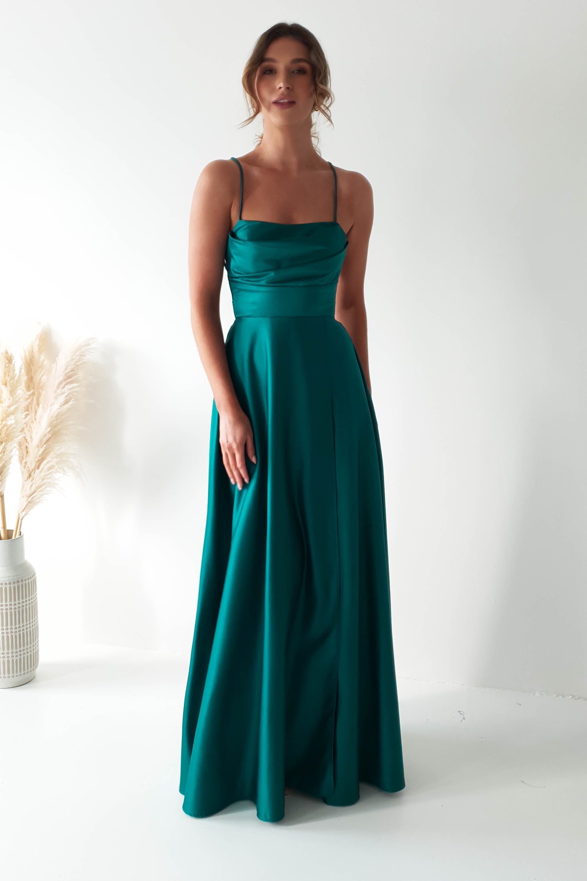 A woman in a teal, floor-length gown with spaghetti straps and a cowl neckline stands against a plain white background. Her hair is styled up, and she is standing beside a vase with pampas grass.