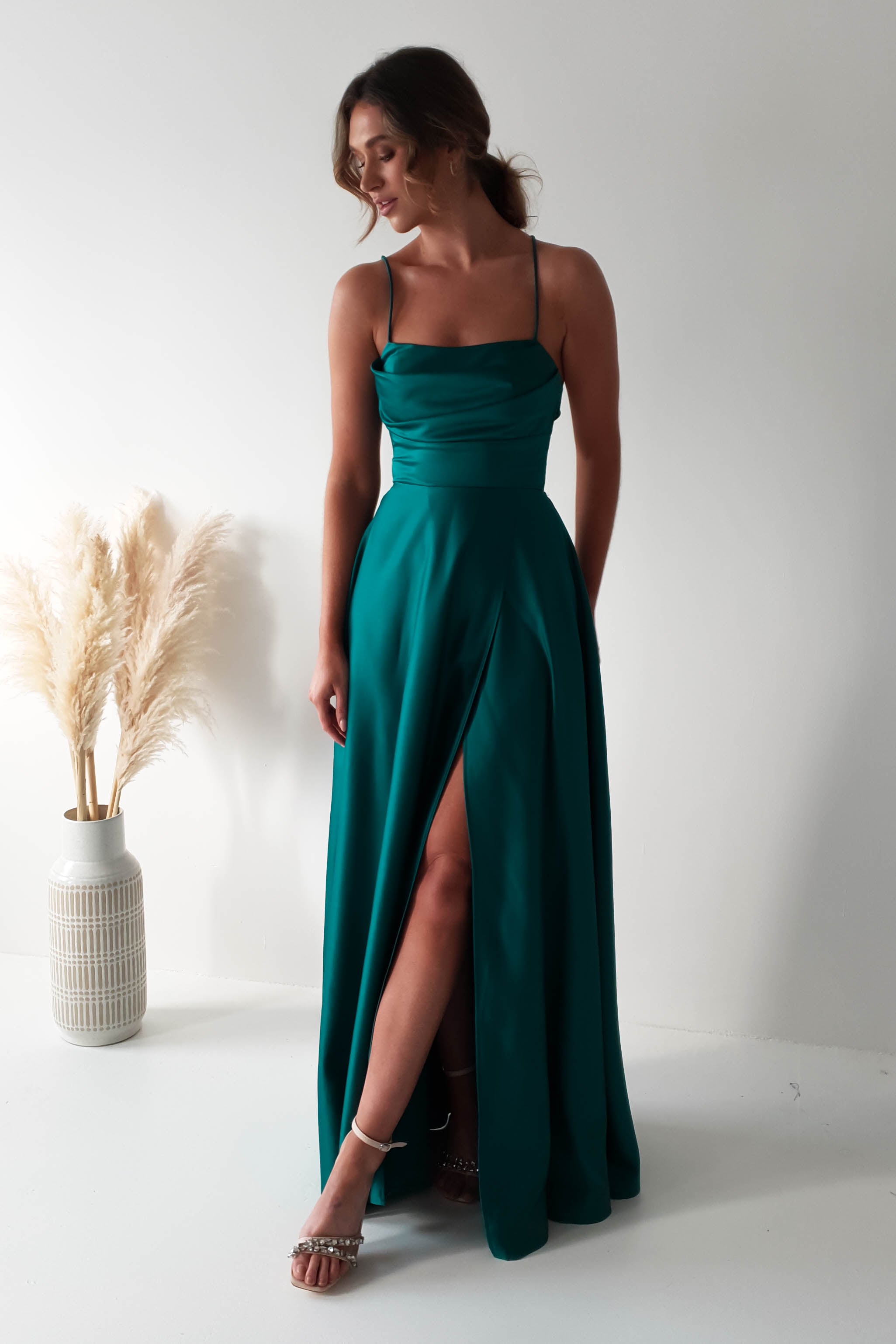 A woman in a teal polyester gown with a cowl neckline elegantly poses in a well-lit room. Her dress features a thigh-high slit, and she complements her look with silver heels. Beside her, a large white vase filled with pampas grass stands against the plain white background.