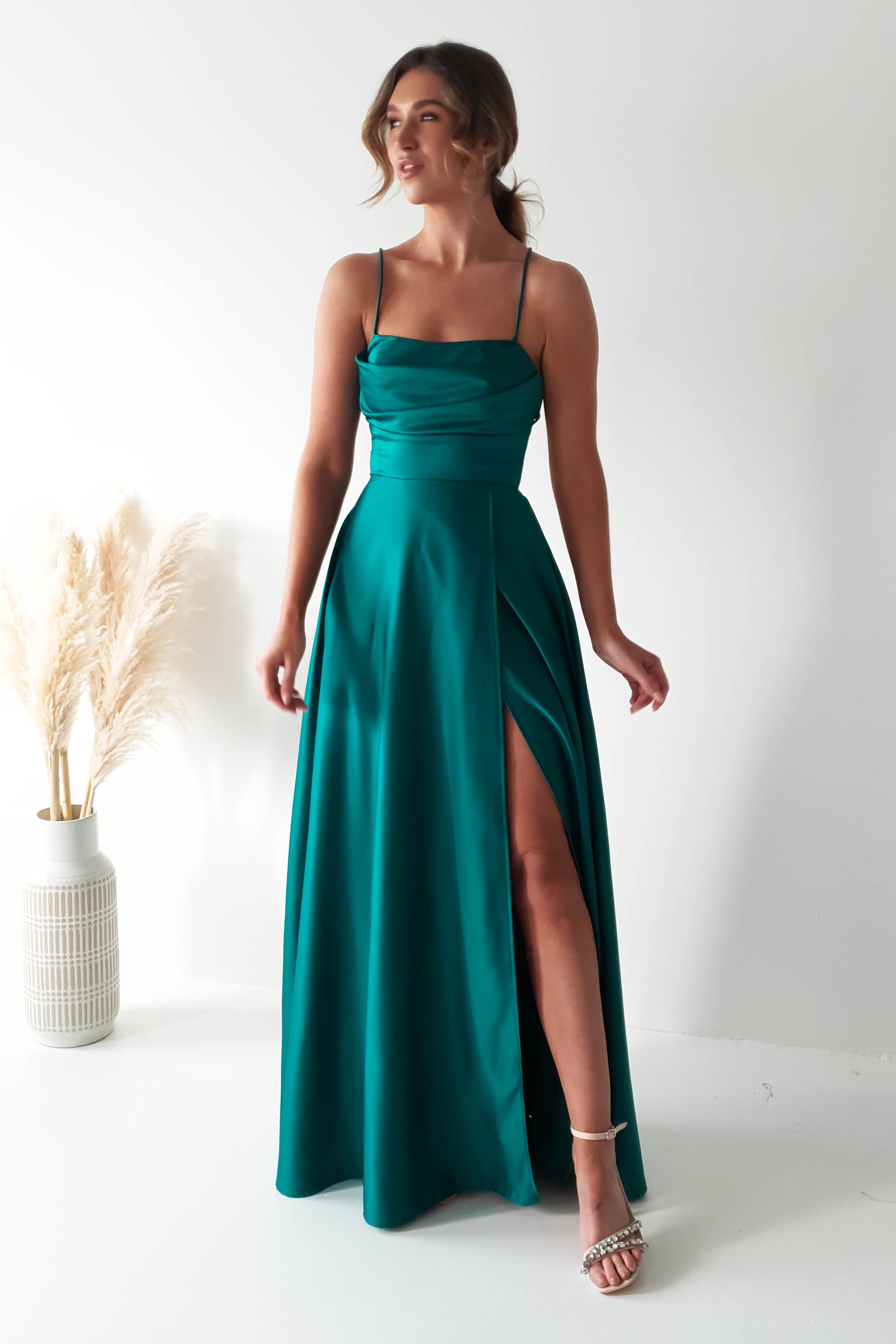 A woman in a teal polyester gown with spaghetti straps, a cowl neckline, and a daring high slit stands in a minimalist room. Her hair falls in loose waves, and she wears high heels. To her left, a vase filled with pampas grass adds to the elegant simplicity of the scene.