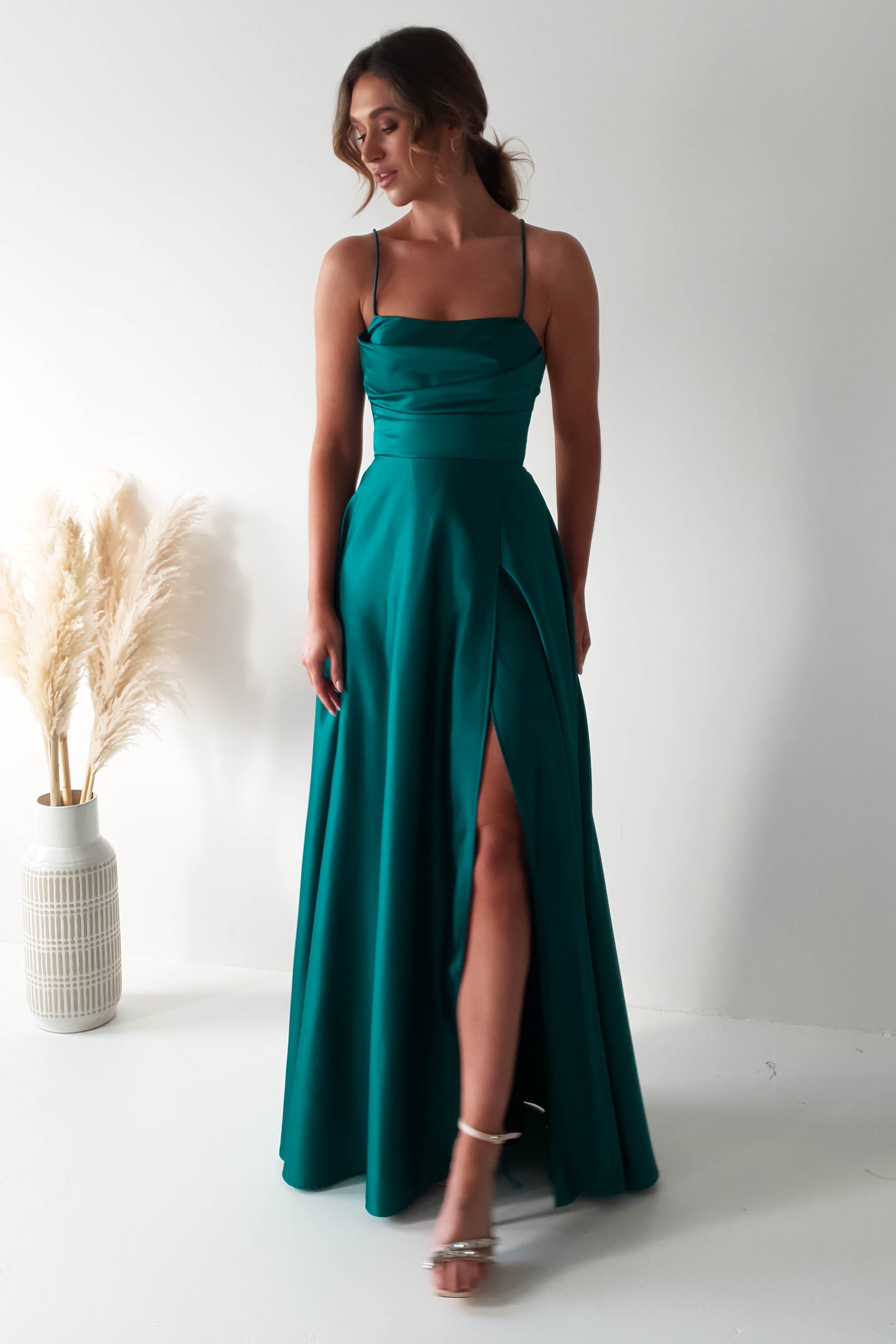 A woman in a teal sleeveless gown, featuring a cowl neckline and a thigh-high slit, stands in a minimalist setting. Her hair is styled back, and she wears strappy heels. The polyester fabric complements the vase with pampas grass decorating the white-walled space.