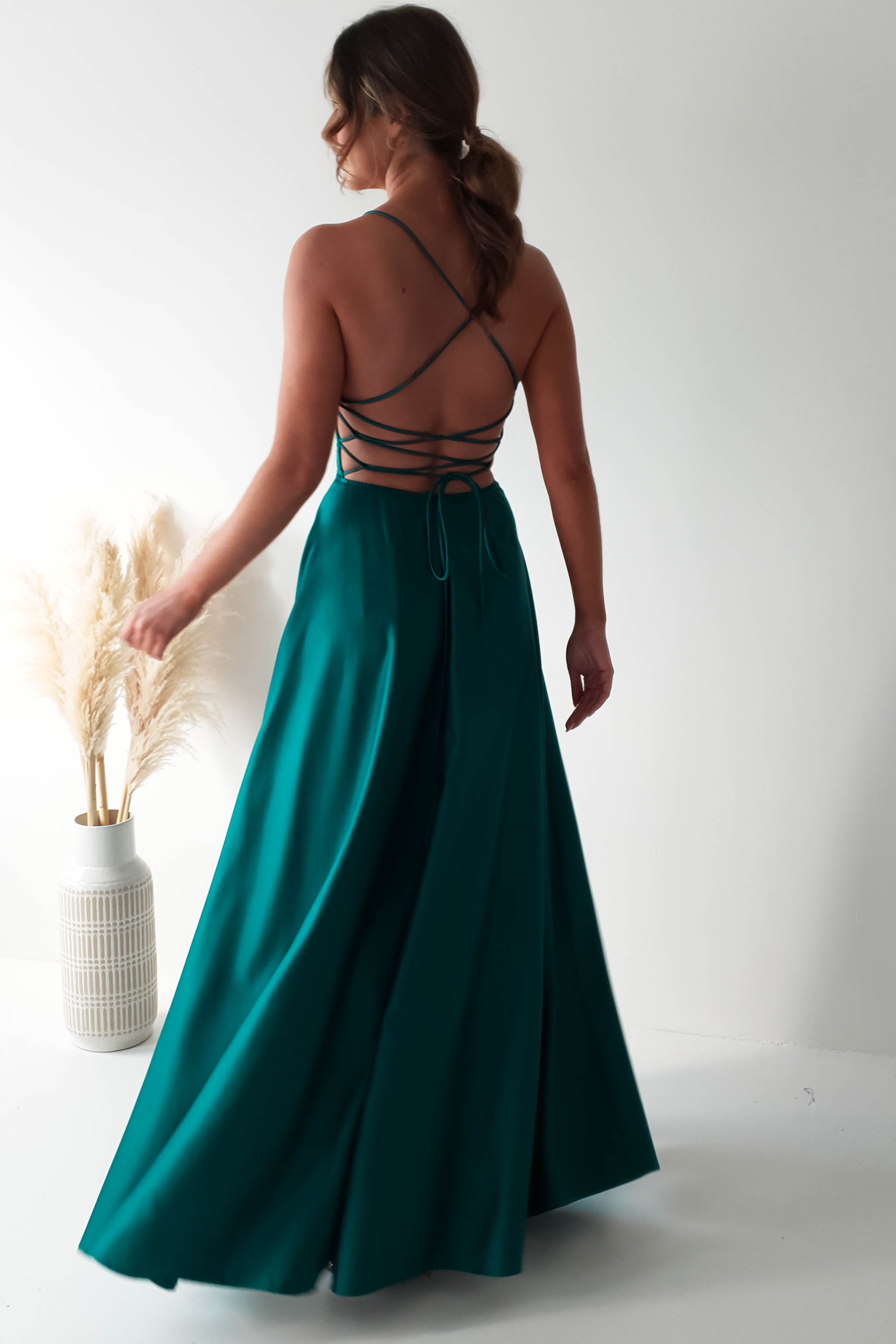A woman in a long, flowing green dress with a cowl neckline stands in a minimalist room against a white wall. The polyester gown features an intricate strappy back design. She is turned slightly, facing a vase with pampas grass on the left.