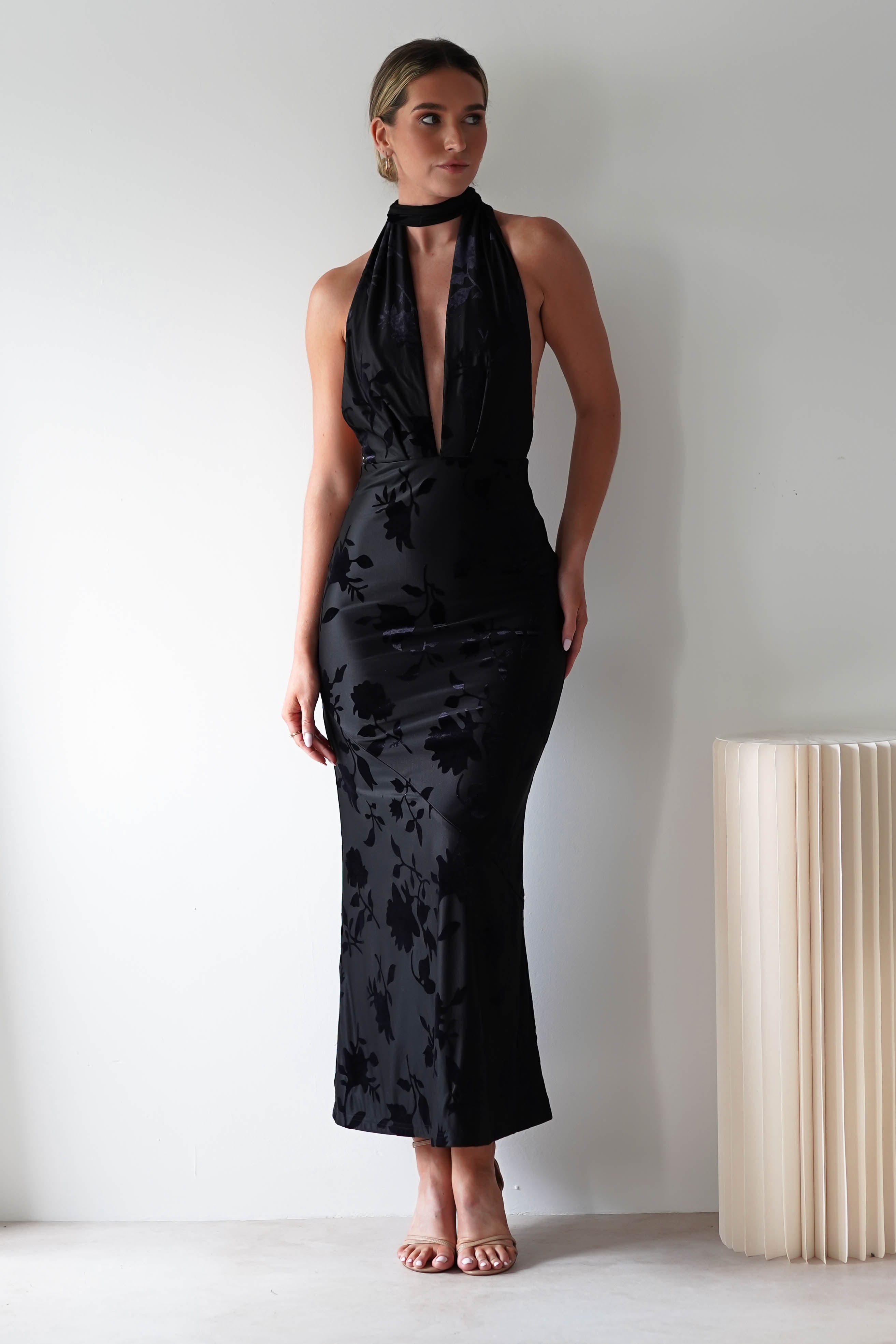 A woman in a sleeveless, black floral dress with a plunging neckline and open back detail balances on her toes beside a minimalist, accordion-style, off-white lamp. The jacquard fabric subtly catches the light as if shes ready to charm as the perfect wedding guest.