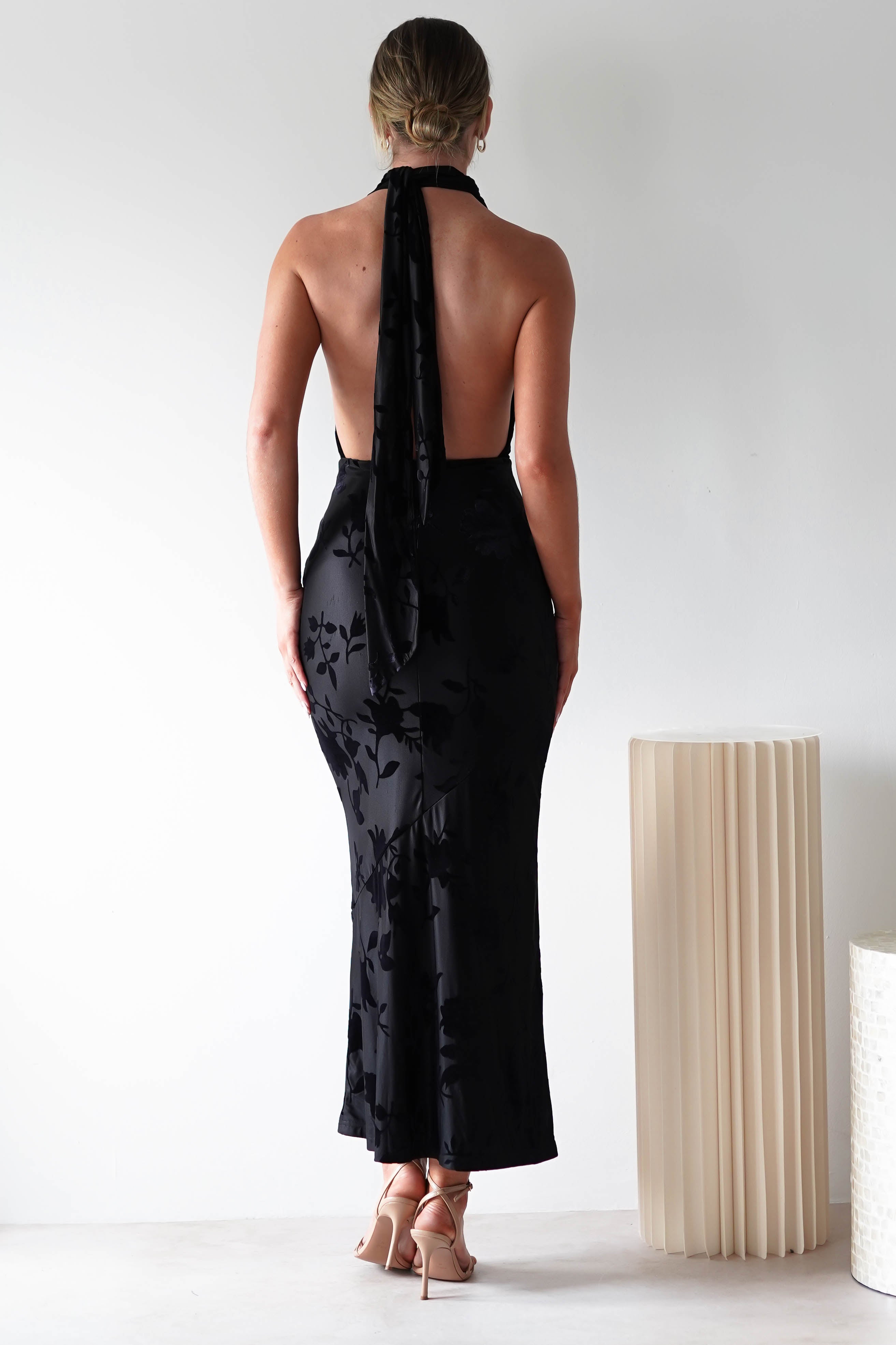A person stands facing away, dressed for a wedding as a guest in a stunning backless black gown with jacquard fabric featuring floral patterns. The halter neck dress, with its open back detail, cascades elegantly to the ankles. They have short hair and are wearing high heels beside a pleated cylindrical decor piece.