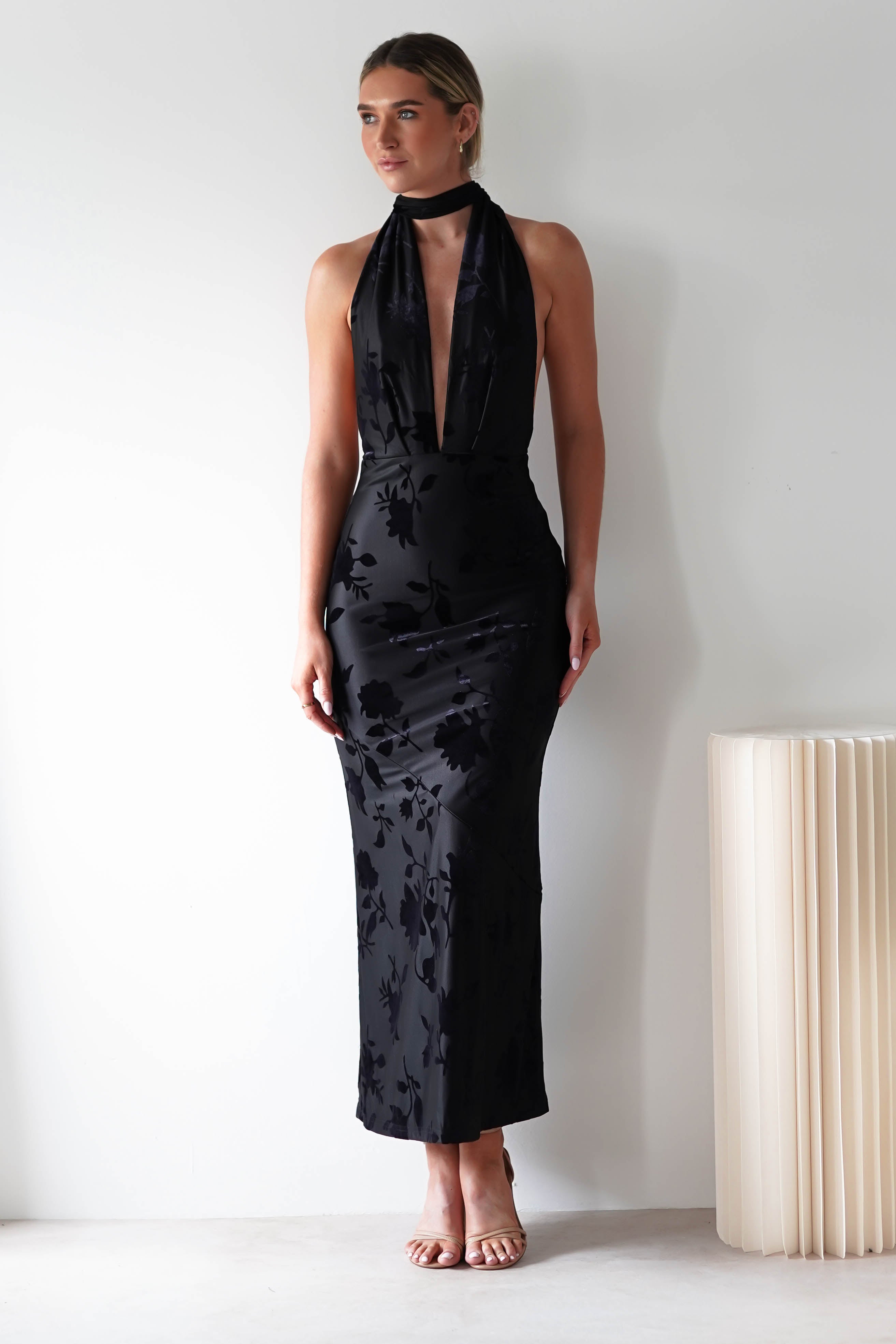 A person stands against a white wall in a sleek, black halter-neck dress with a deep V-neckline and open back detail, perfect for a wedding guest. The floor-length dress features floral patterns in jacquard fabric. They pair it with strappy heels and have their hair pulled back beside a ribbed, cream-colored decorative piece.