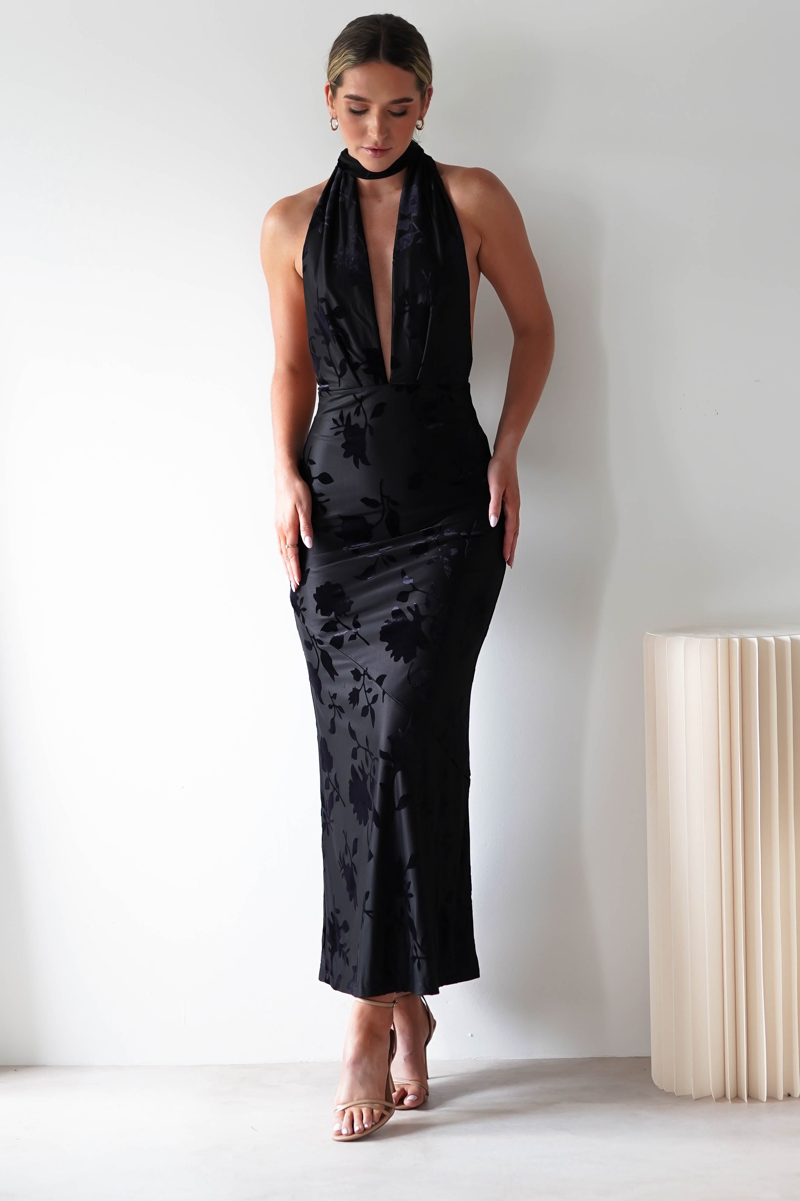 A person stands against a white wall in a sleeveless, deep V-neck, floor-length black dress featuring a subtle floral pattern on luxurious jacquard fabric. With minimal accessories and nude heels, they exude elegance fit for a wedding guest. A beige, pleated object is partially visible on the right.