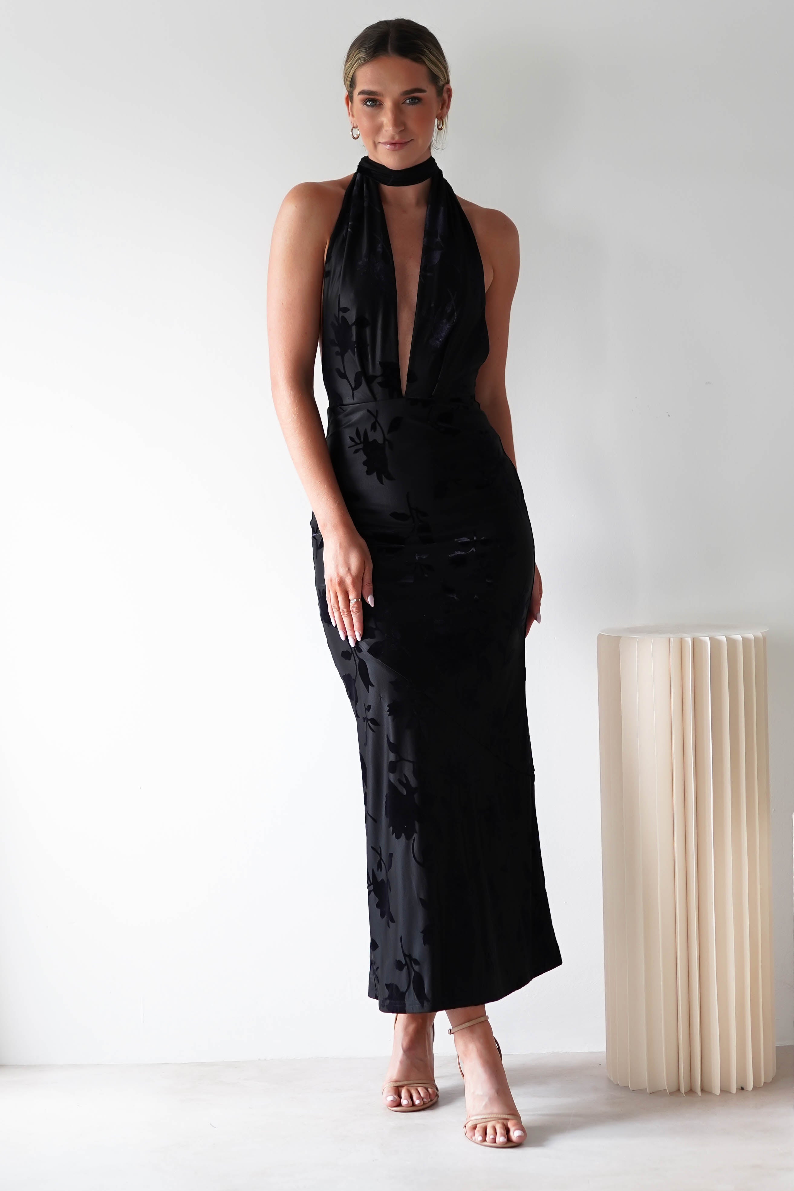 A person stands against a plain white background wearing a sleeveless, floor-length, halter-neck black dress with a plunging neckline and an open back detail. With light-colored hair tied back and nude high heels, they pose next to a beige, cylindrical pedestal.
