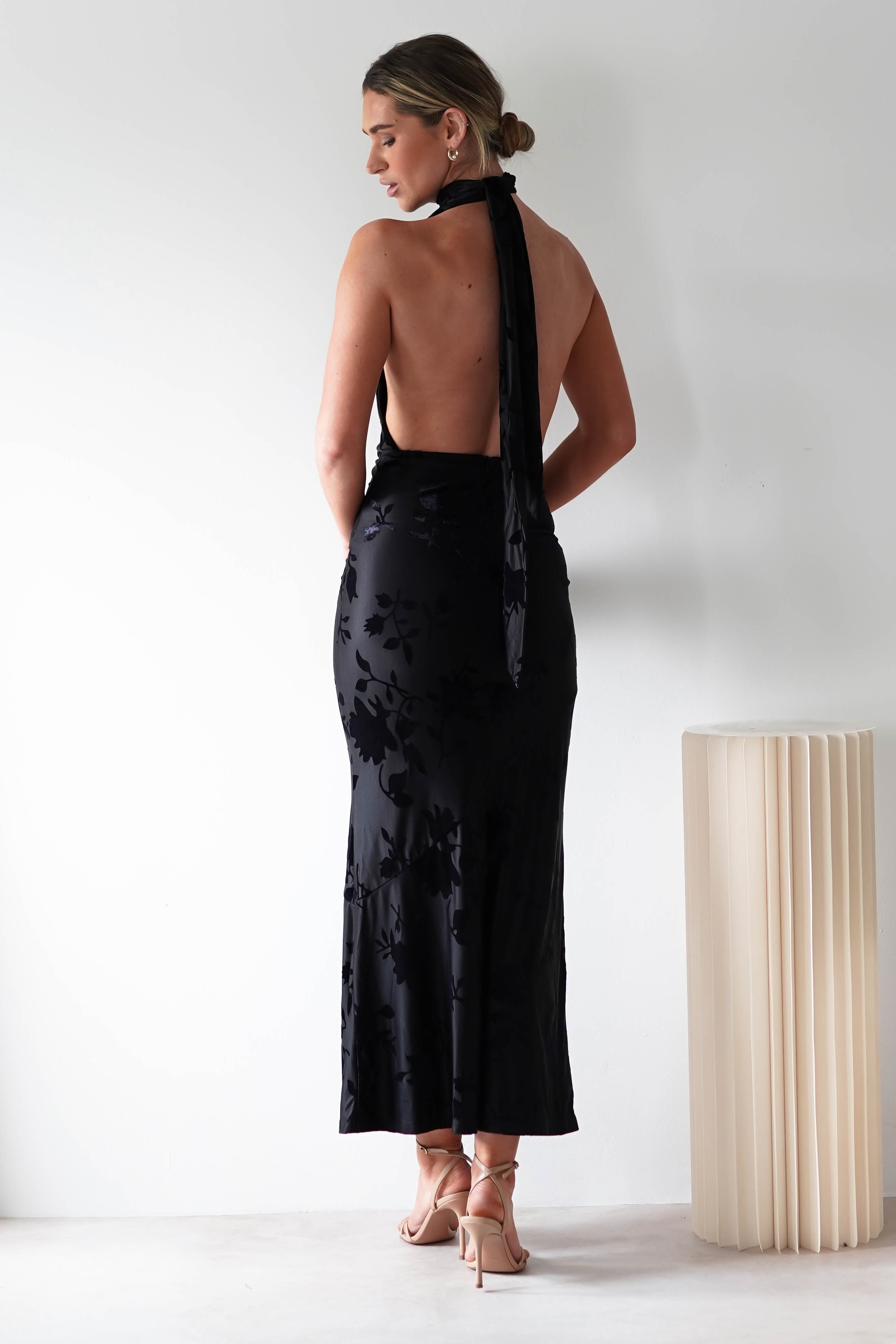 A person stands facing away, wearing a backless black dress with floral patterns and a halter neckline tied at the back. Made from elegant Jacquard fabric, it complements their hoop earrings and high-heeled sandals perfectly. A tall, pleated light fixture is nearby.