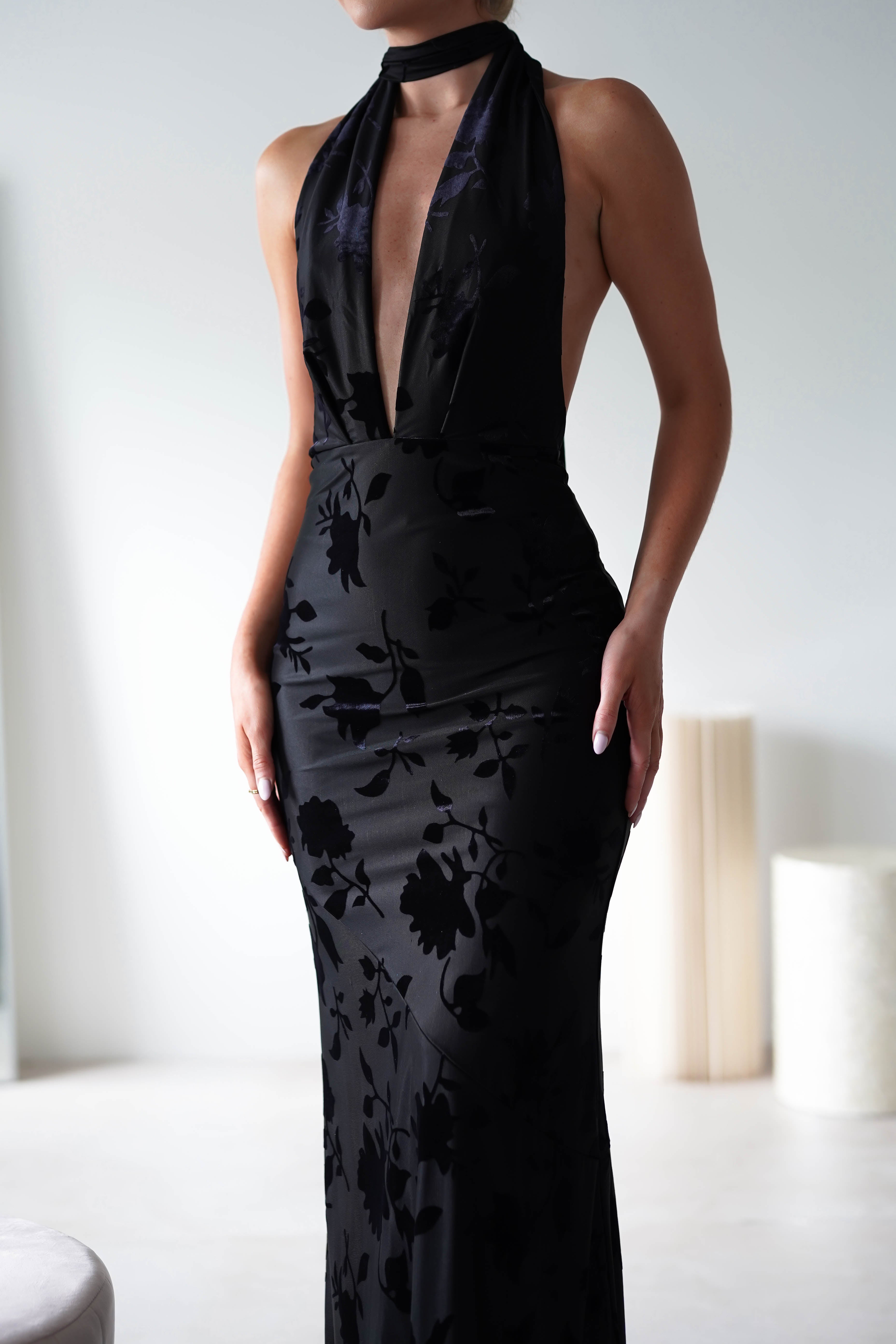 A person wearing an elegant long black dress with a deep neckline and floral pattern, crafted from luxurious Jacquard fabric. The form-fitting dress features an open back detail and is paired with a black choker. The simple indoor setting has light-colored furniture, perfect for a wedding guest appearance.