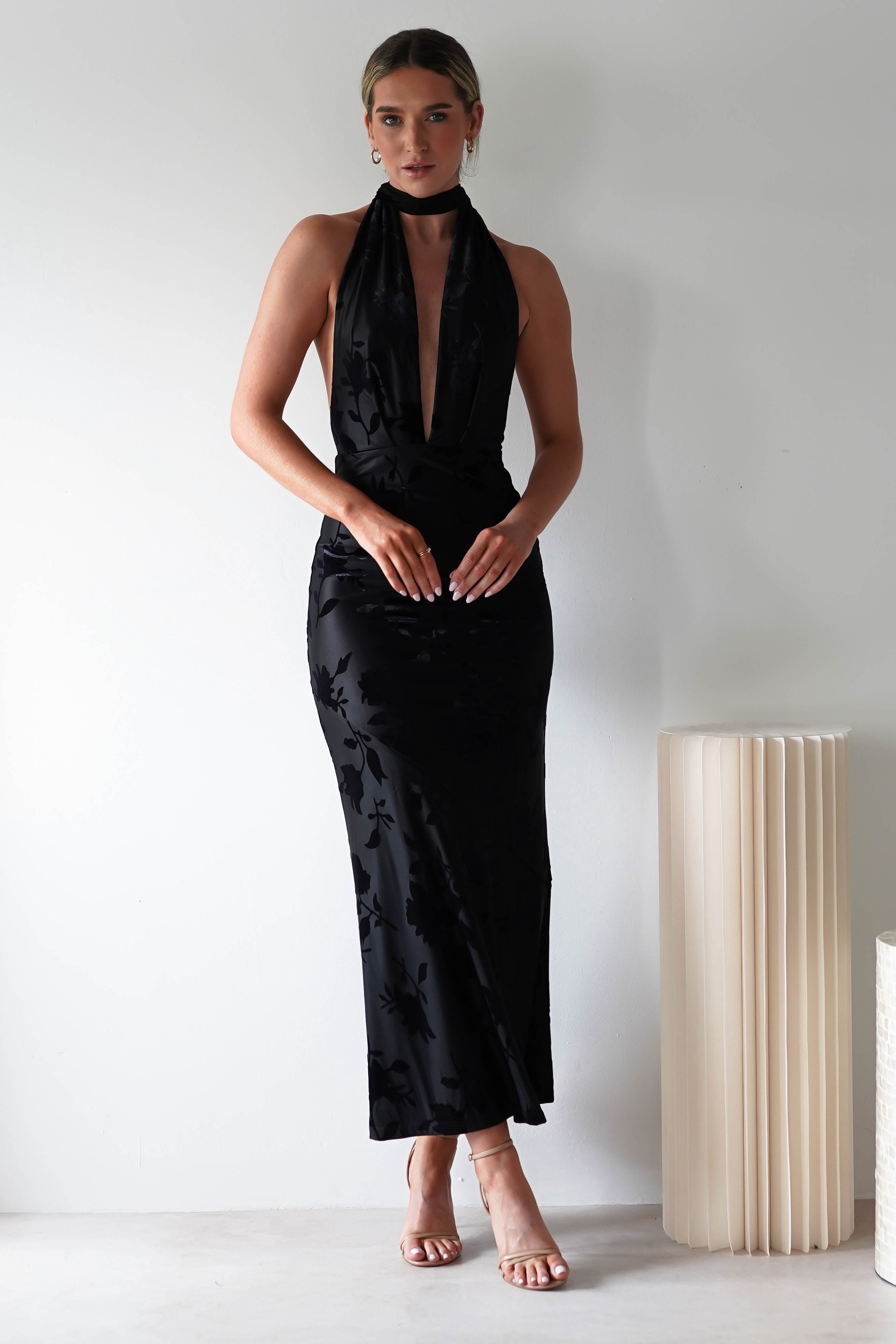 A person stands against a white wall in a long, elegant black halter neck dress made of jacquard fabric, featuring a deep V-neckline and floral patterns. The open back detail adds allure as they gently clasp their hands in front. A tall, cylindrical lamp quietly accents the scene.
