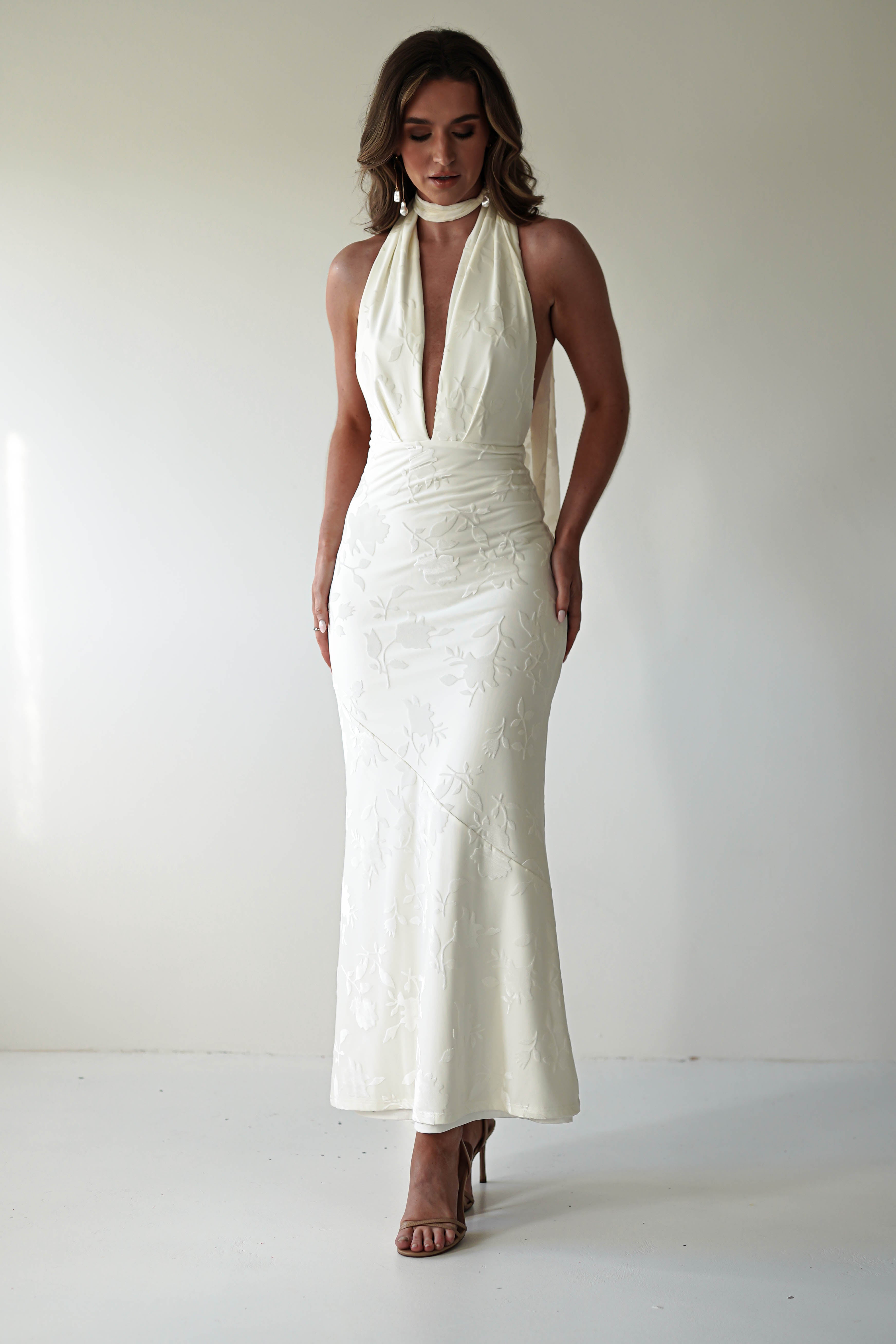 The woman, exuding elegance in her jacquard fabric gown with a deep V-neckline and glamour neck-tie, poses against a plain backdrop. Her long hair cascades down as high-heeled sandals complete the look, while subtle floral patterns add a touch of grace.