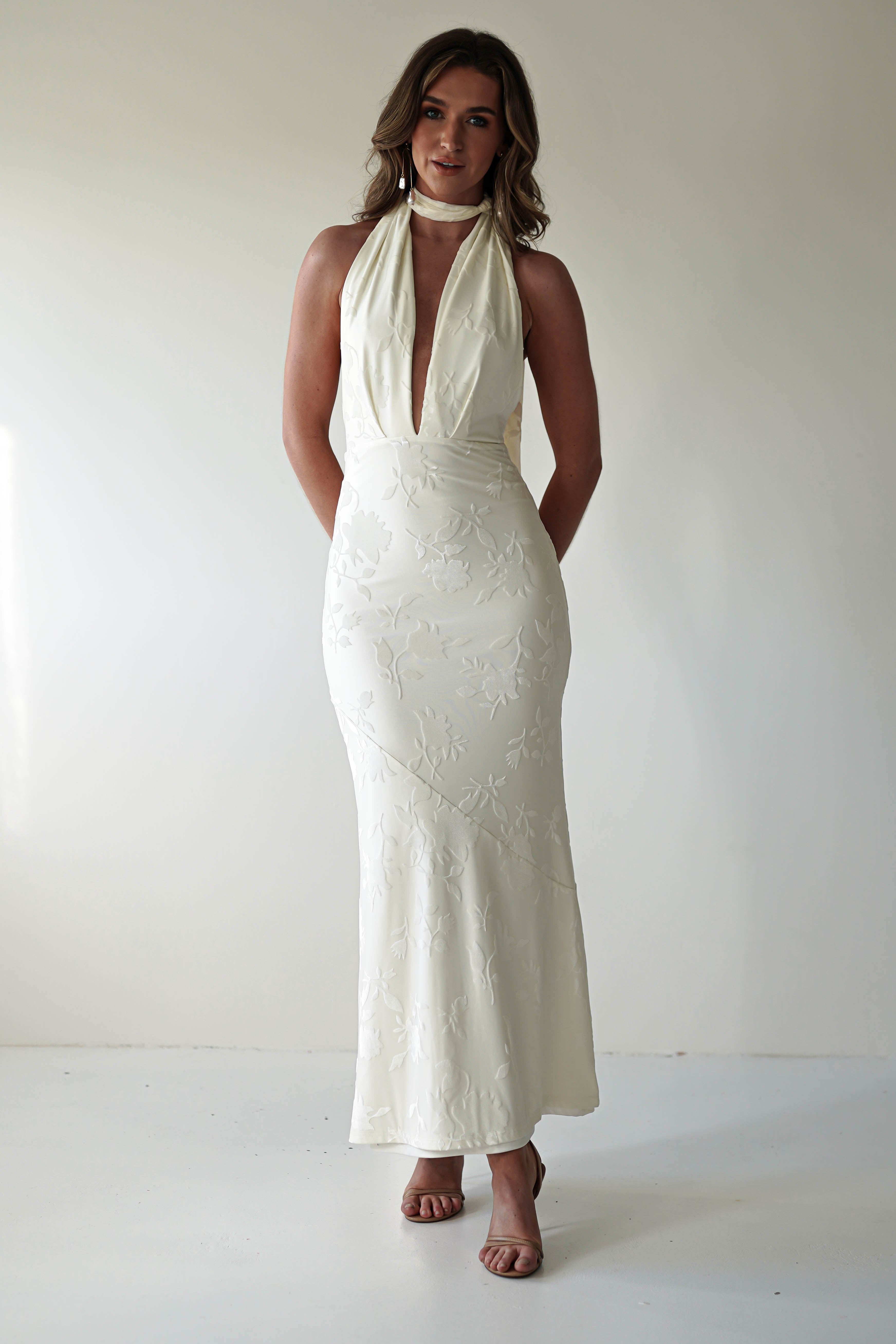 A person stands against a plain background, wearing a patterned, sleeveless white dress crafted from jacquard fabric with a plunging neckline and halter style. They have long hair and are wearing high-heeled sandals, perfect for elegant events or christenings.