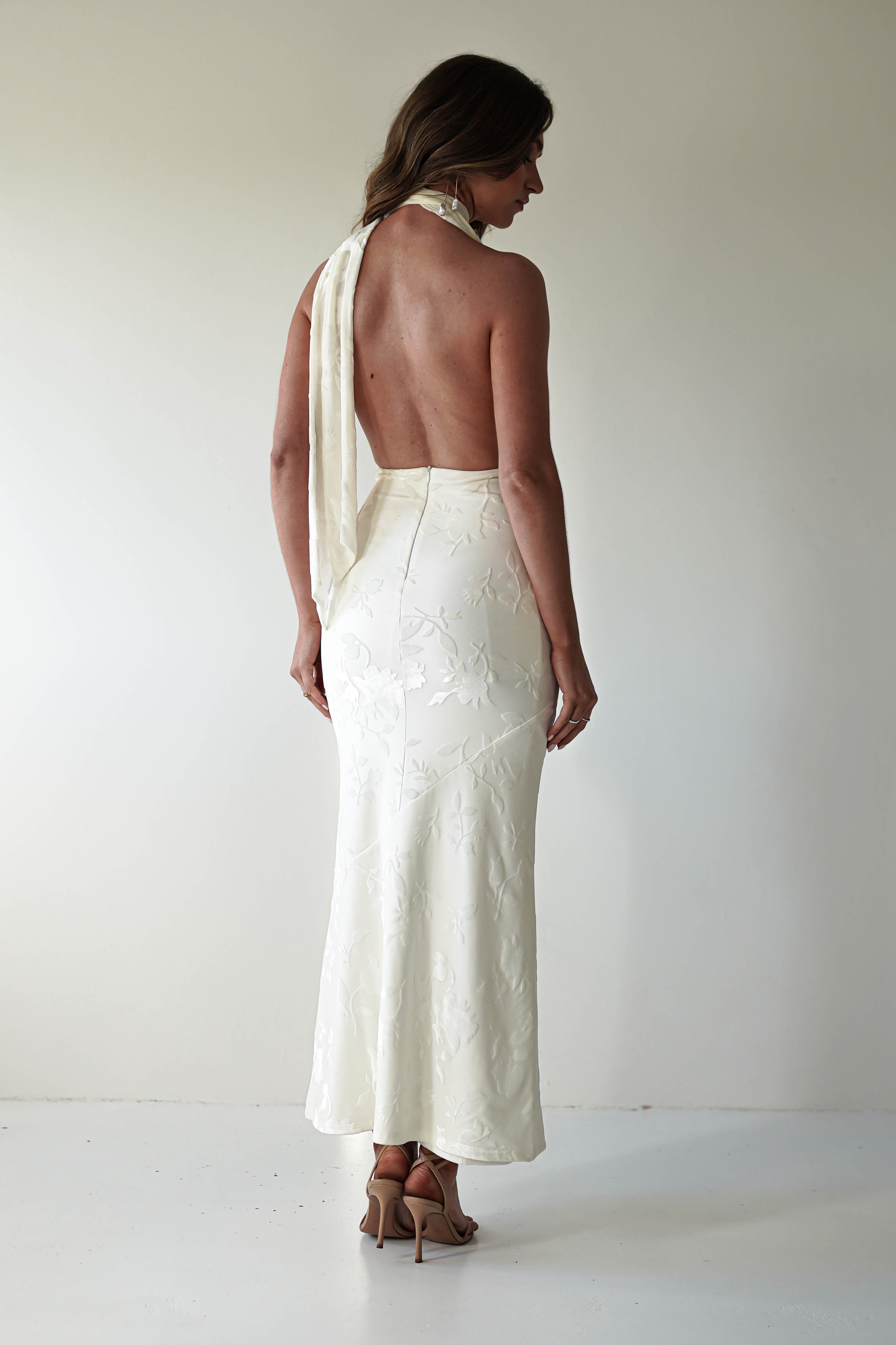A woman in a backless, floral-patterned ivory dress made of jacquard fabric, tied at the neck in a glamourous fashion, stands facing away. She is wearing high heels against a plain, light-colored wall and floor.