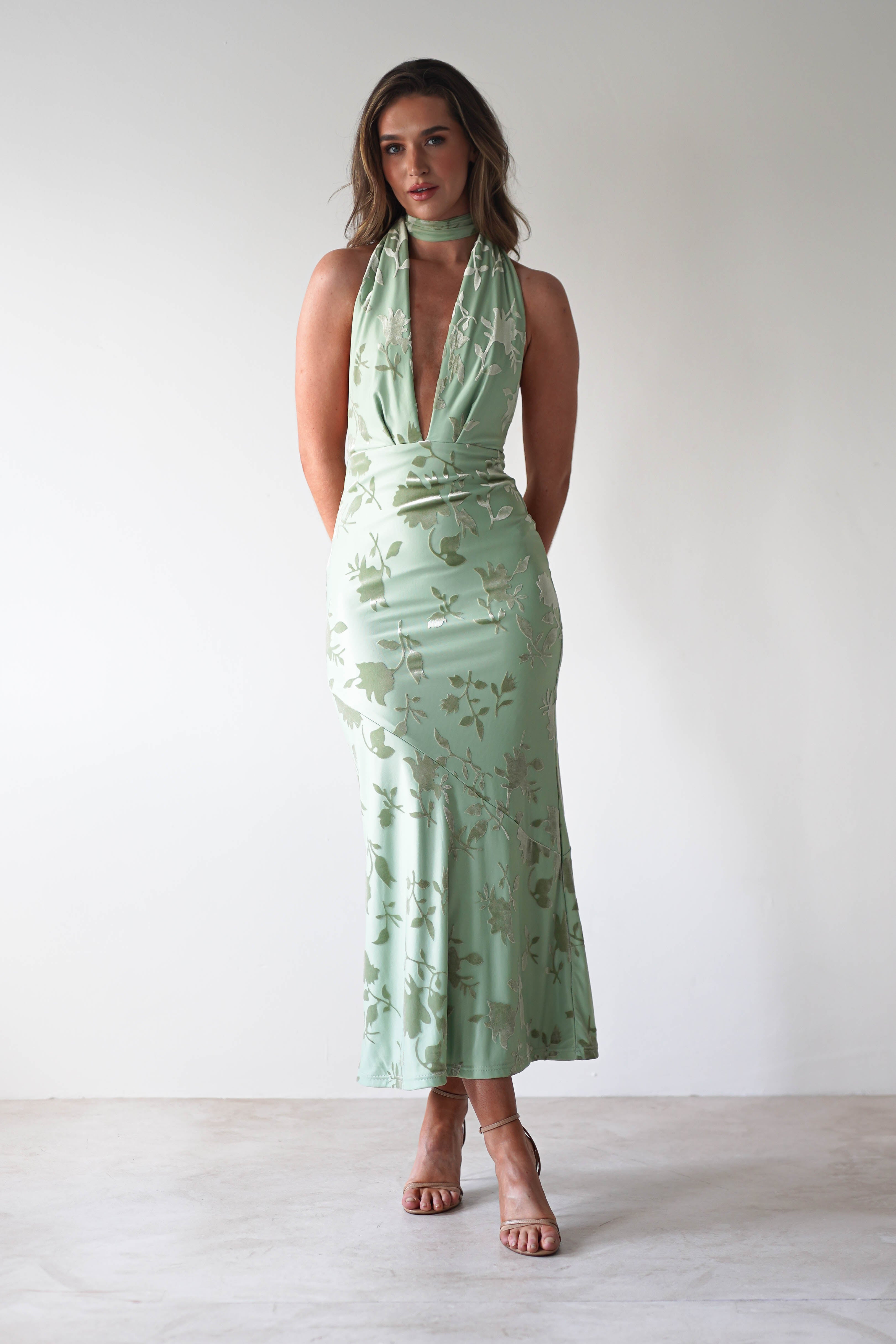 A person stands against a plain white background, wearing a sleeveless light green dress with a floral jacquard fabric pattern and a deep V-neck. The adjustable tie straps add elegance. With long hair cascading down and strappy heels, theyre impeccably dressed as the perfect wedding guest, hands clasped behind their back.