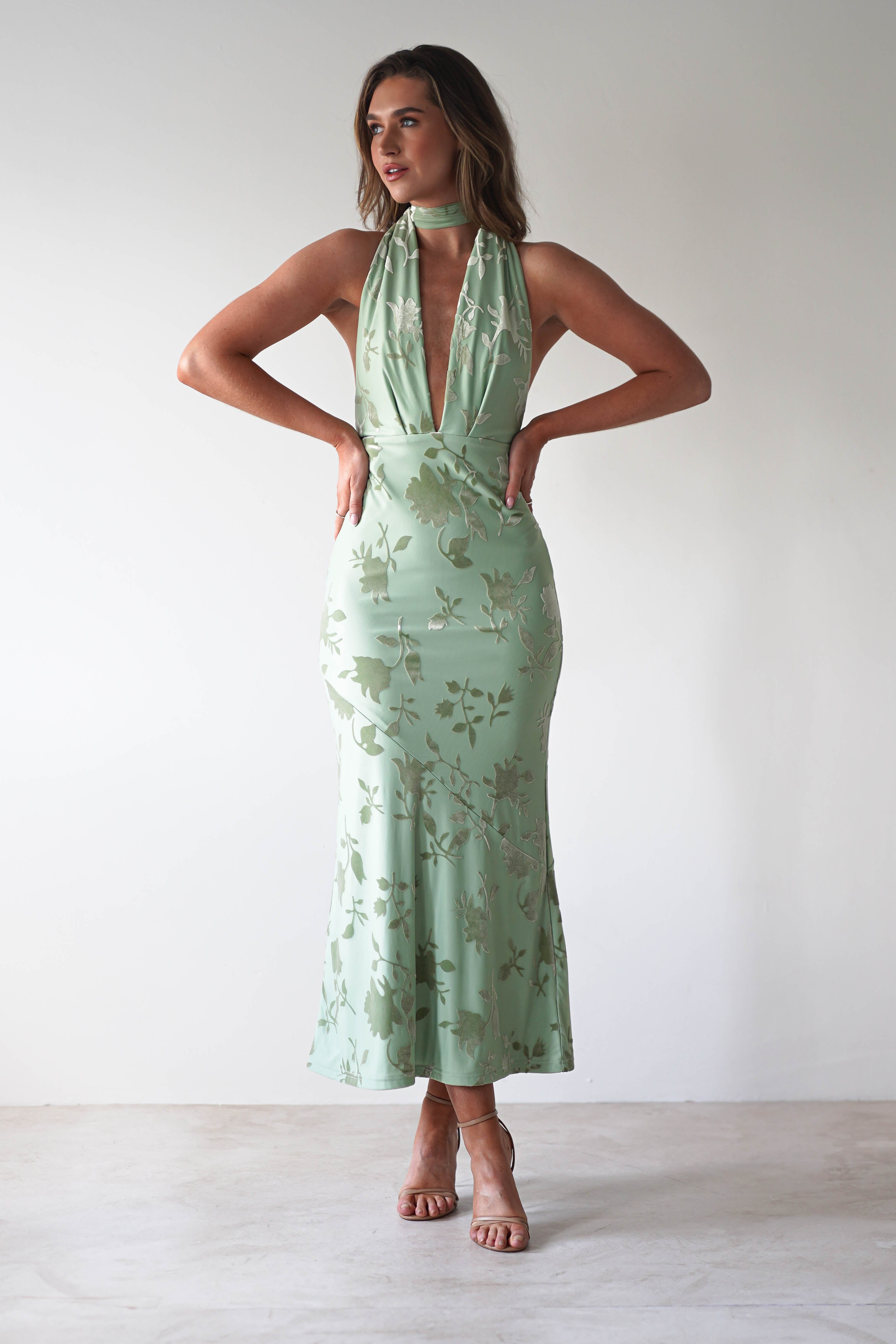 The woman, dressed as a stylish wedding guest, flaunts a mint green halter-neck dress with an elegant floral pattern. The deep neckline and fitted silhouette complement her look, accented by adjustable tie straps. Beige high heels complete the ensemble against a plain white backdrop.