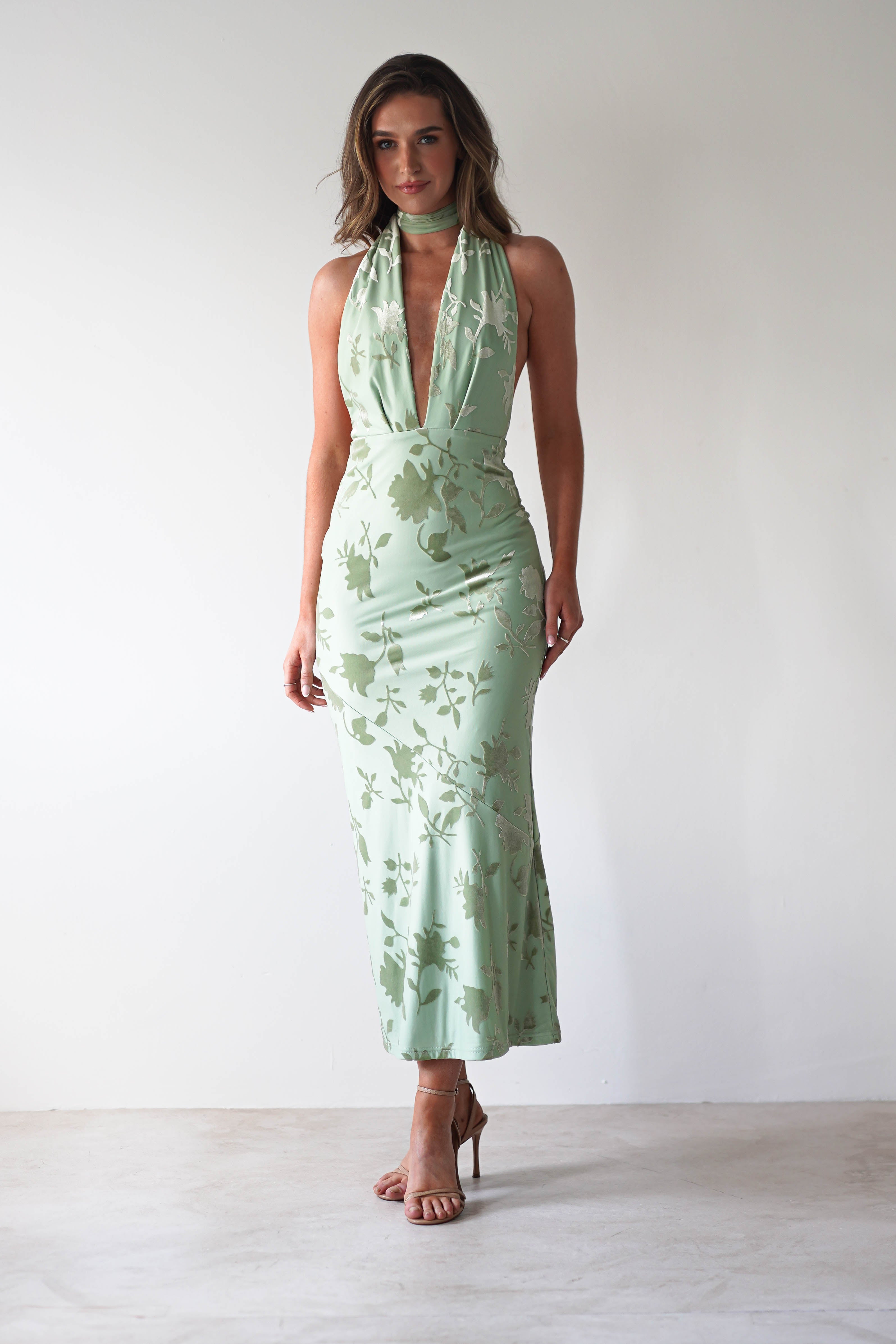 A woman stands against a plain white background, wearing a light green jacquard fabric dress with a floral pattern and plunging neckline. The dress features an adjustable tie strap, perfect for any wedding guest look. She has long hair and wears high-heeled sandals, her hands relaxed at her sides.