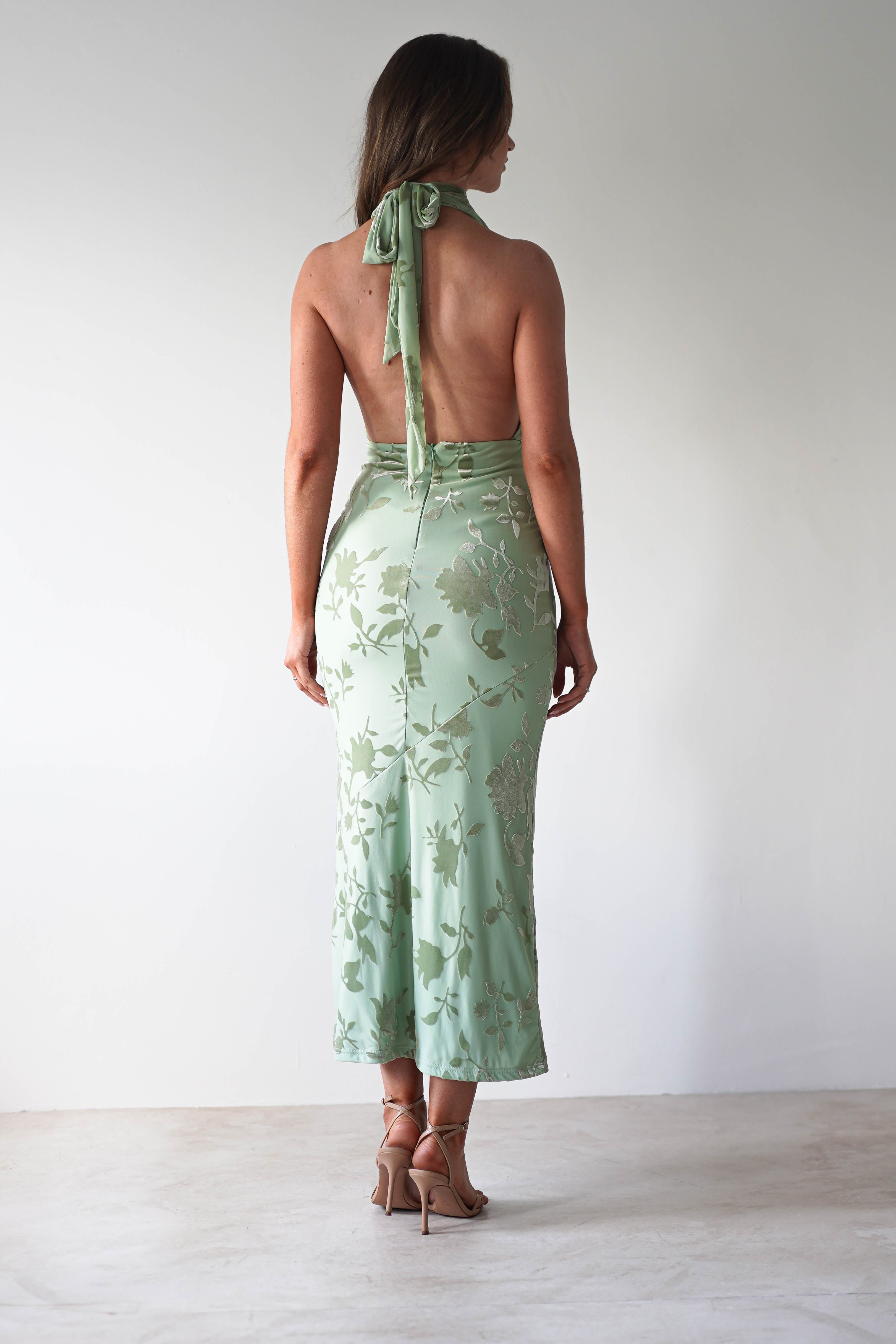 A woman in a light green halter dress made of jacquard fabric stands with her back to the camera. The dress features a floral pattern and an open back, tied with adjustable straps at the neck. She wears tan high heels on a light-colored floor against a plain white background.