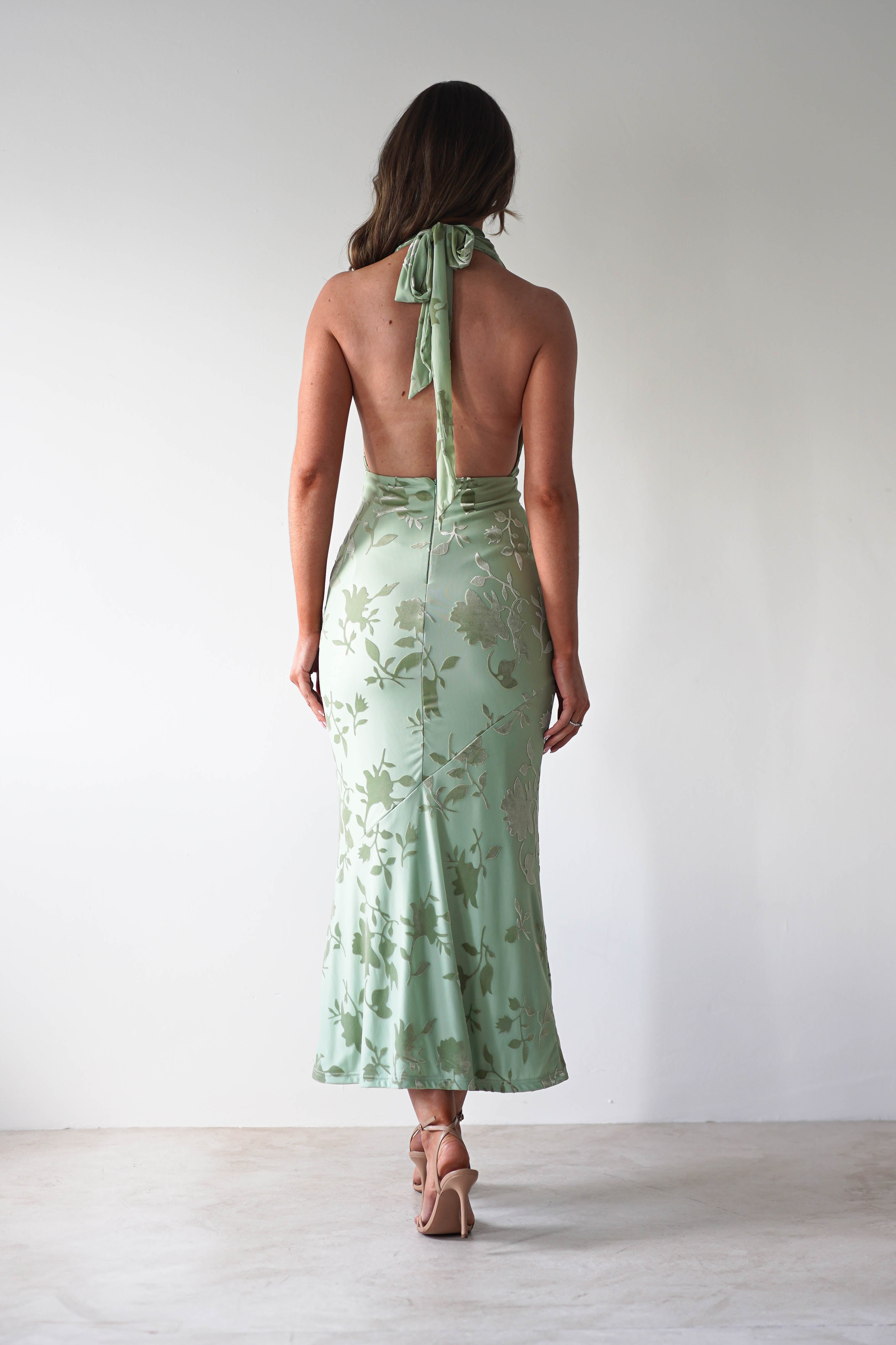 A woman stands facing away, wearing a light green, backless dress with floral patterns, perfect for a wedding guest. Crafted from jacquard fabric, the dress ties at the neck and flows to a calf-length hem. She completes her look with beige high heels against a plain background.