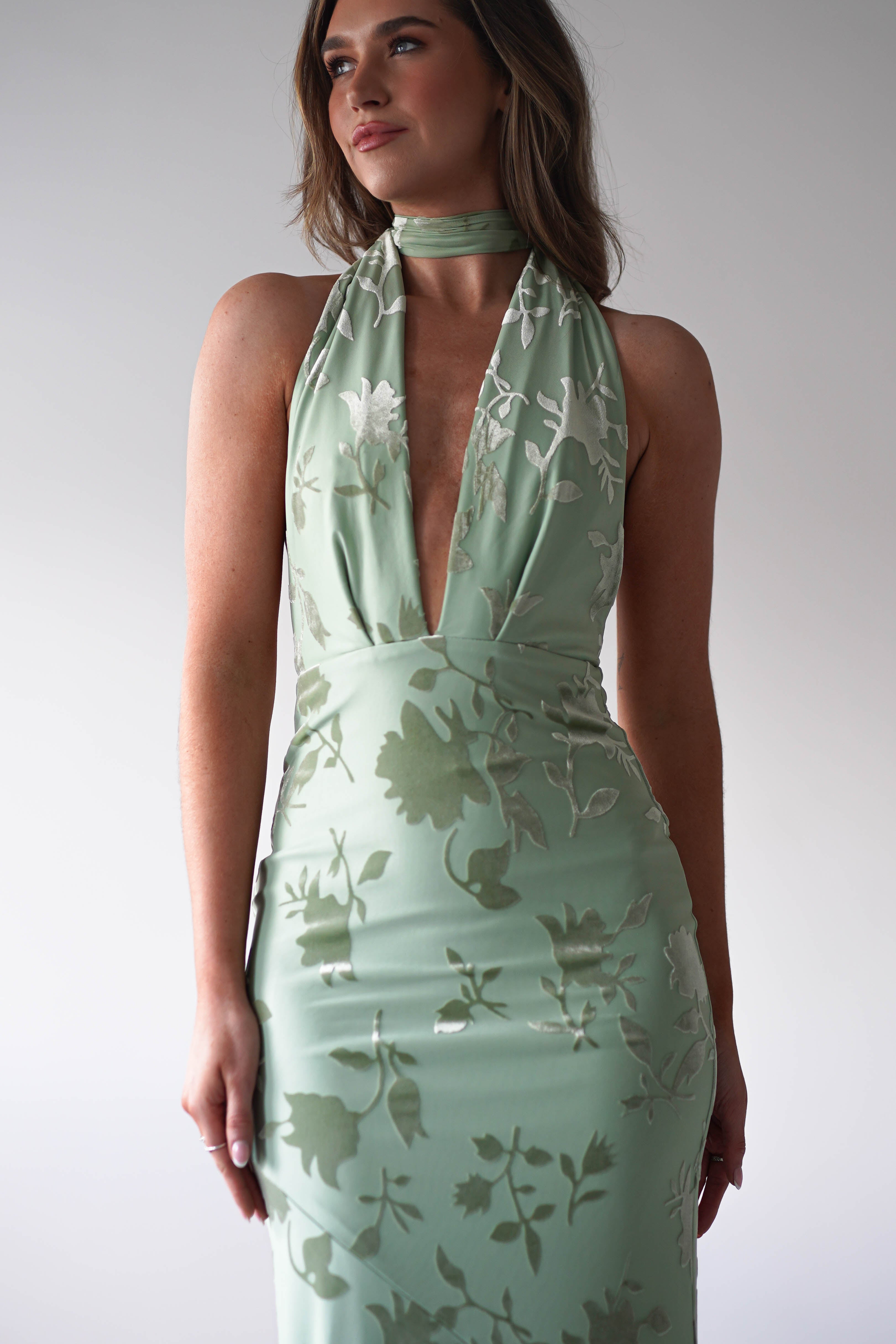 A woman in a light green, floral-patterned halter dress with a deep V-neckline and adjustable tie straps stands against a plain background, looking slightly upward. Her hair is styled loosely, and her expression is calm, perfect for a wedding guest ensemble.