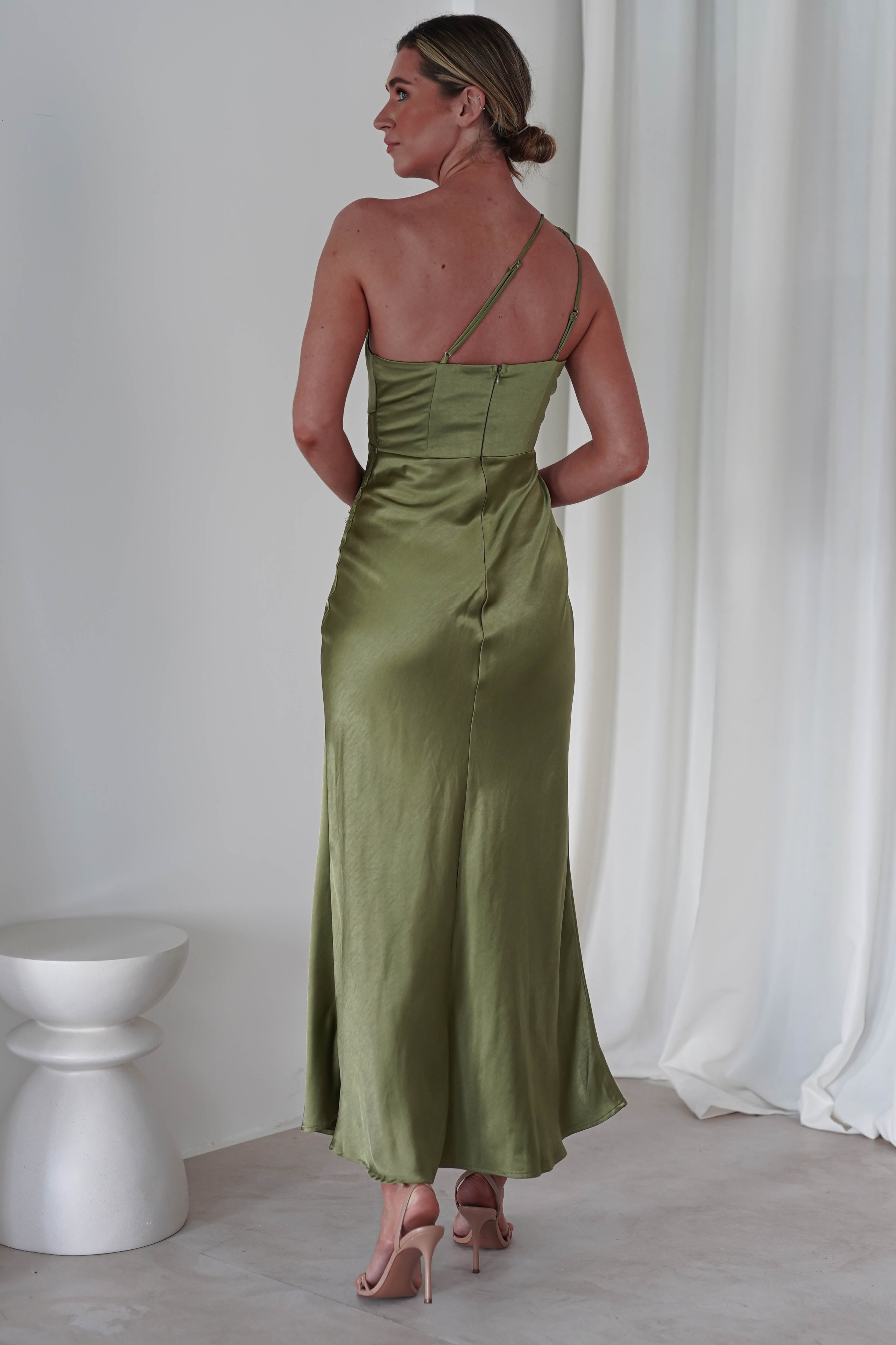 A woman stands with her back to the camera, wearing an elegant satin maxi dress in green with thin straps. Her hair is styled up, and she is in a room with white curtains and a white stool, the perfect ensemble for a sophisticated wedding guest. High heels complete her look.