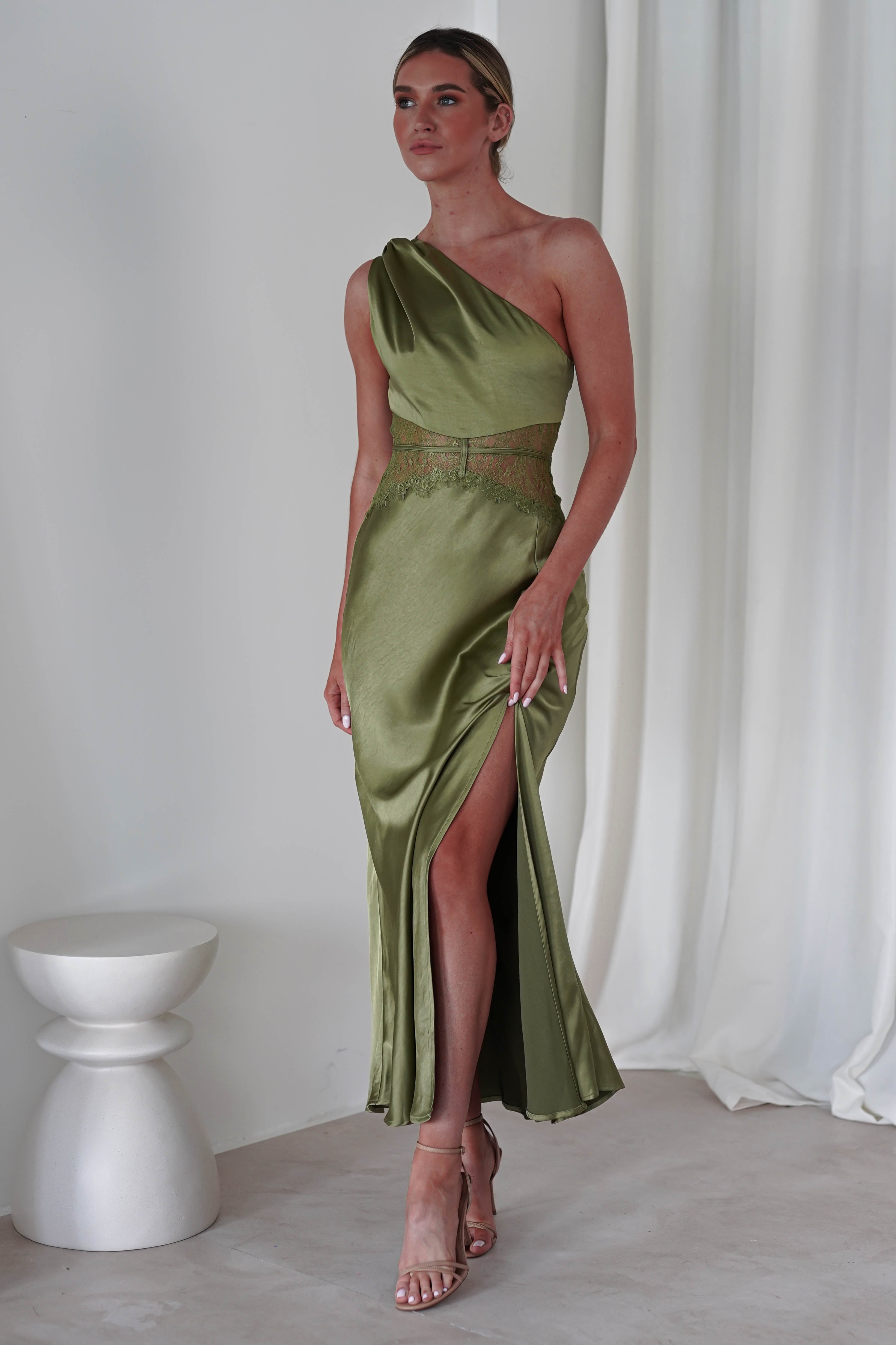 A person in a green, one-shoulder satin maxi dress with a high slit stands indoors. The dress features embroidered lace detailing at the waist. They wear strappy heels and have their hair tied back. White curtains and a small white table are in the background, perfect for a wedding guest look.