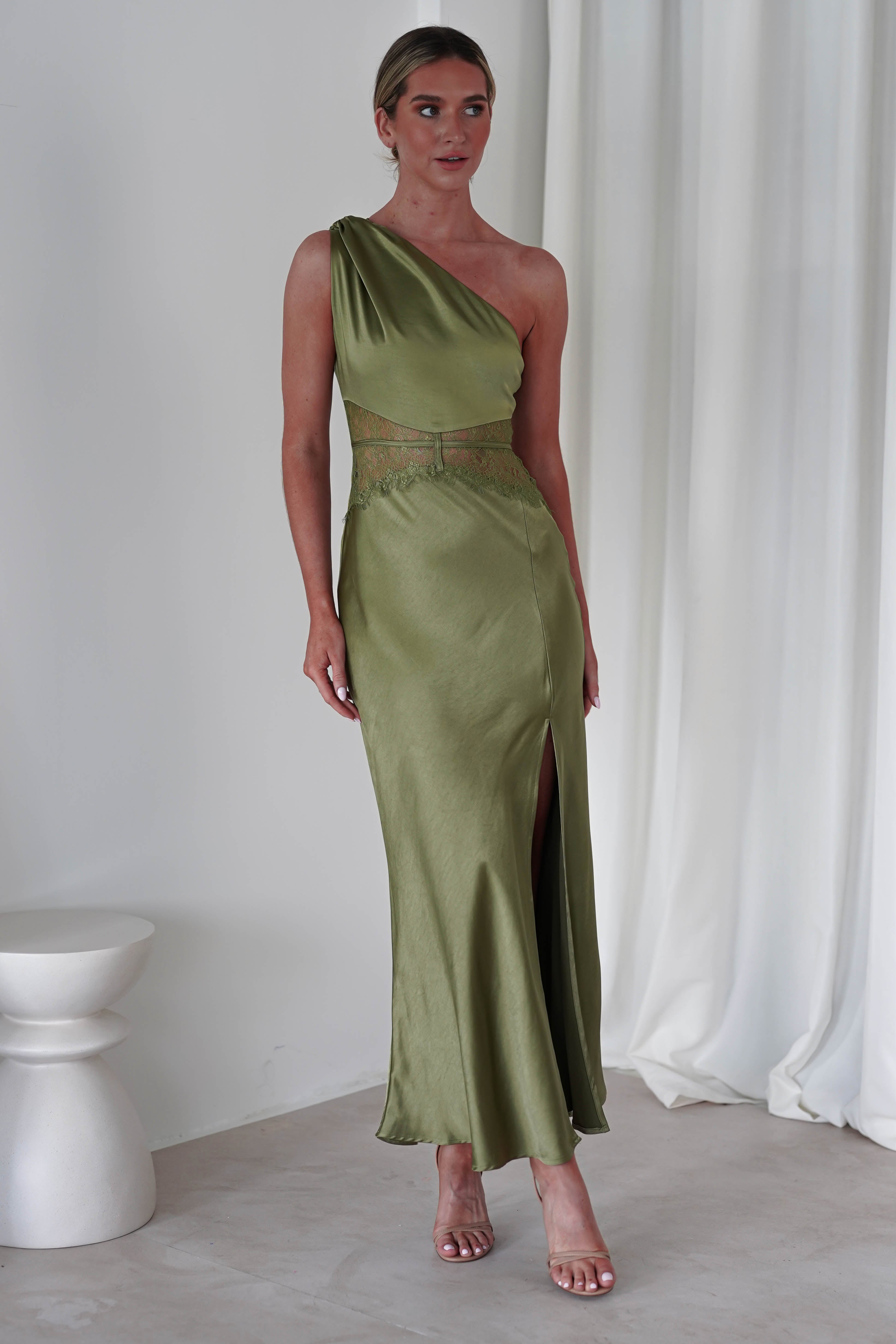 A person stands wearing a sleek, olive green satin maxi dress with a thigh-high slit. The dress features an embroidered midriff and delicate cut-out detail at the waist. They stand in a minimalist setting with soft white drapes and a ceramic stool, perfect for a wedding guest.
