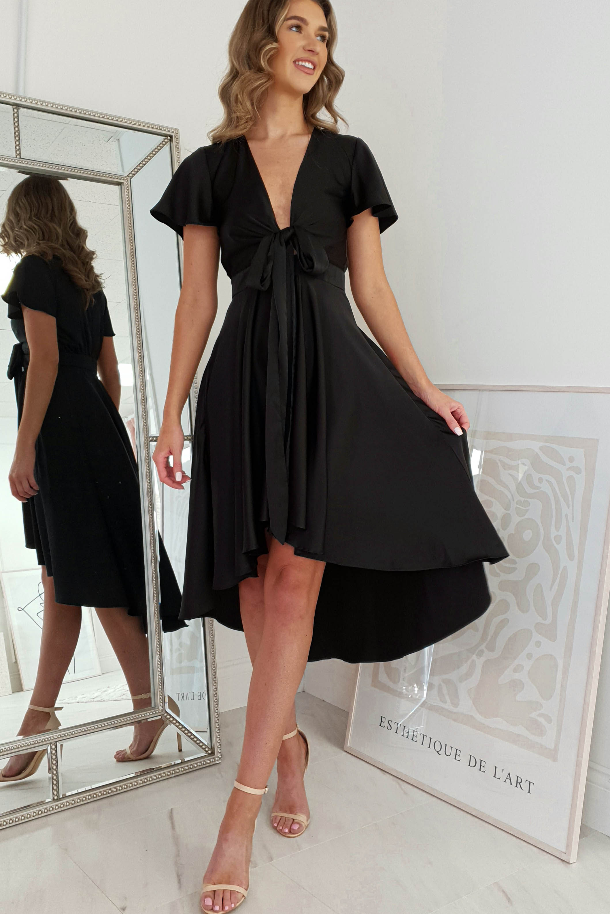 A woman in a stylish black V-neck dress stands in a bright room, gently holding the skirt out to the sides. She smiles while glancing to her left. A large mirror and framed artwork with abstract patterns adorn the background.