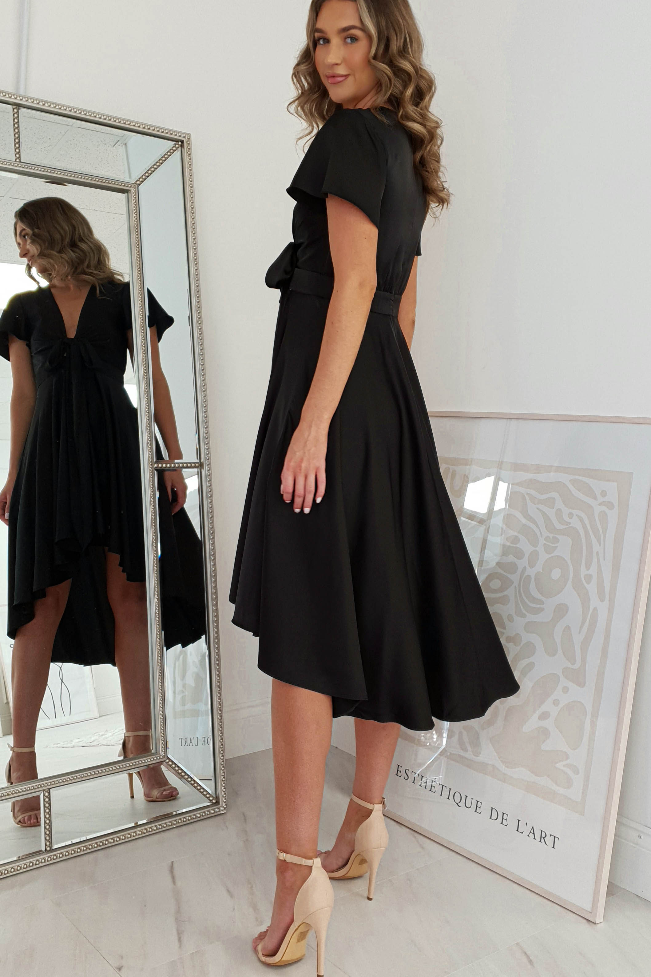 A woman in a stylish black dress with nude heels poses in front of a mirror. The V-neck dress features a cinched waist and a high-low hem. A framed abstract print leans against the wall on the light wood floor, enhancing her chic appearance.