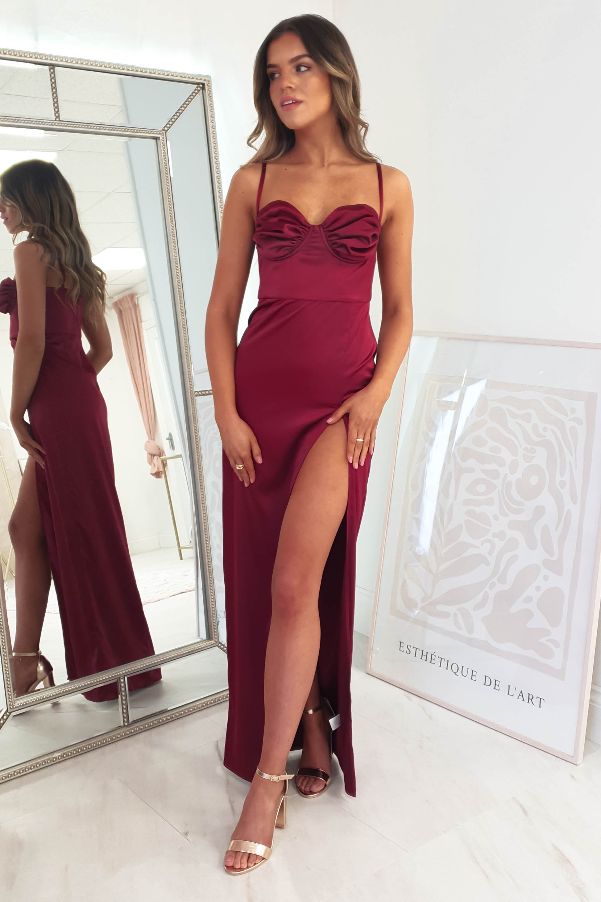A woman in a red dress with adjustable straps and a high thigh split stands by a mirror, wearing gold heels. A framed abstract artwork is partially visible on the floor. The setting exudes a bright, soft, and elegant ambiance, perfect for a formal event.