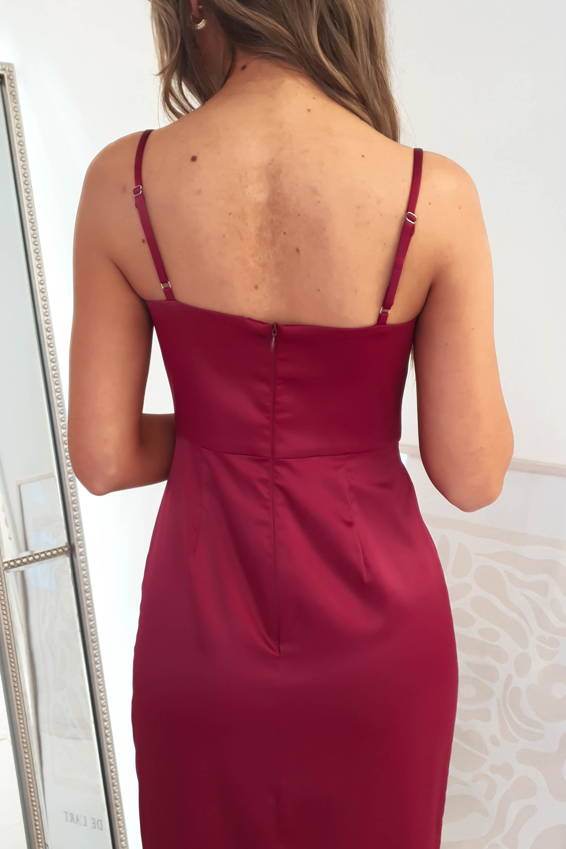 A woman with long hair is wearing a red sleeveless dress with adjustable straps, viewed from the back. She stands in front of a mirror, where the reflection hints at a formal event and part of the rooms decor.