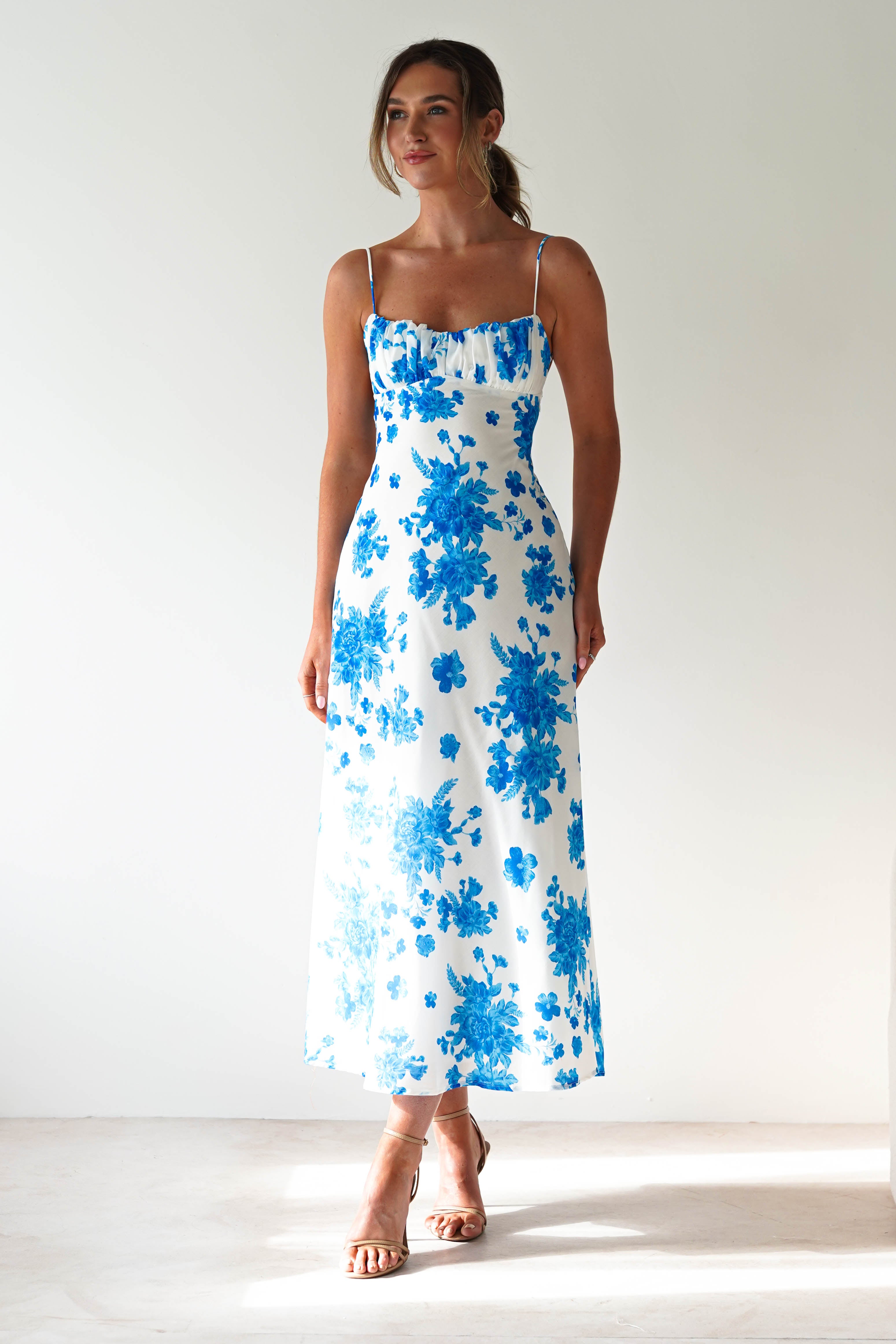 A woman stands wearing a blue midaxi dress with spaghetti straps, blue floral prints, and beige heeled sandals, perfect for a wedding guest look against a plain light background.