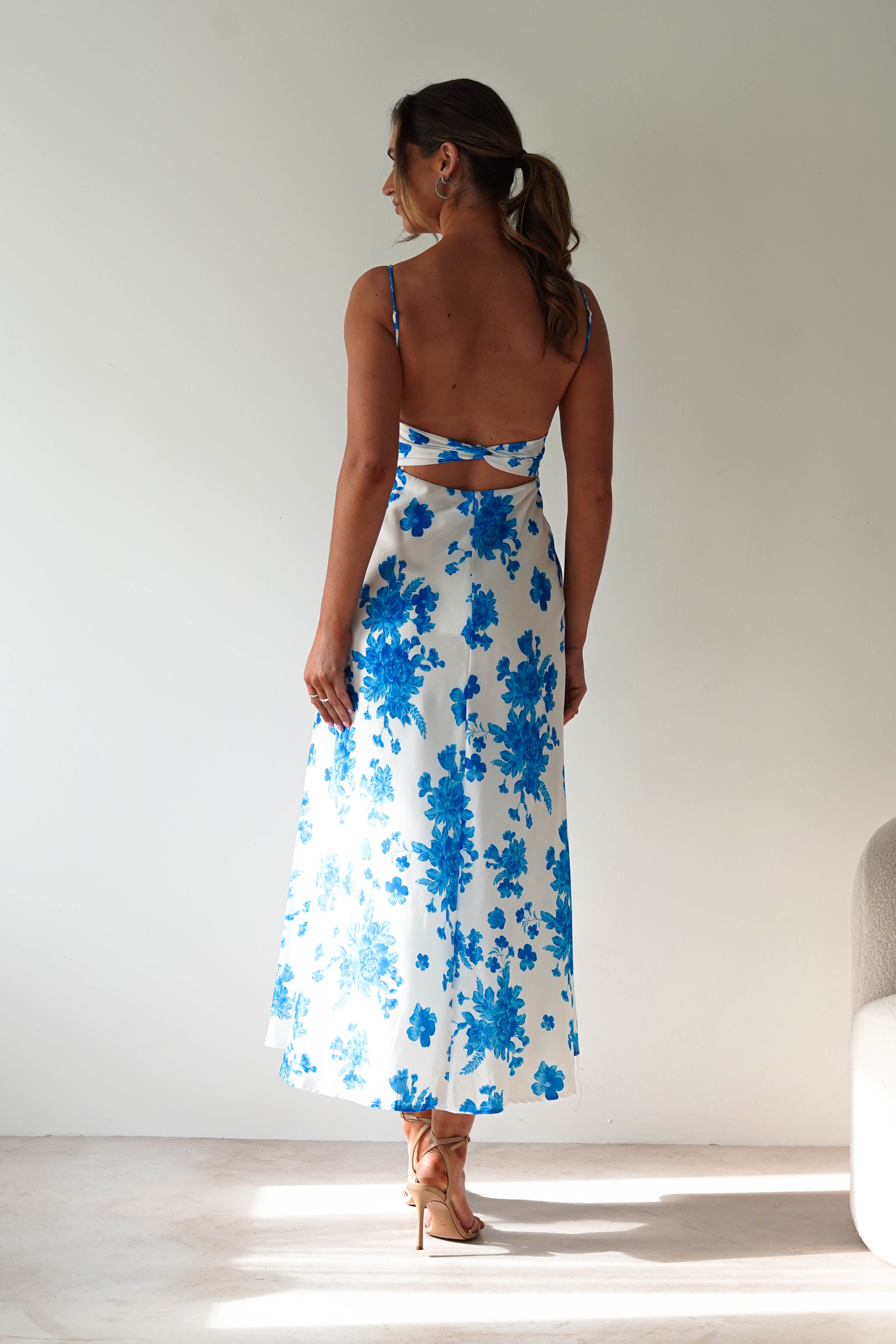 A woman stands indoors facing away, wearing a blue midaxi dress with white floral patterns and spaghetti straps. Her hair is tied in a low ponytail, and she pairs the look with beige heels. Light streams in from the left—a perfect wedding guest style.