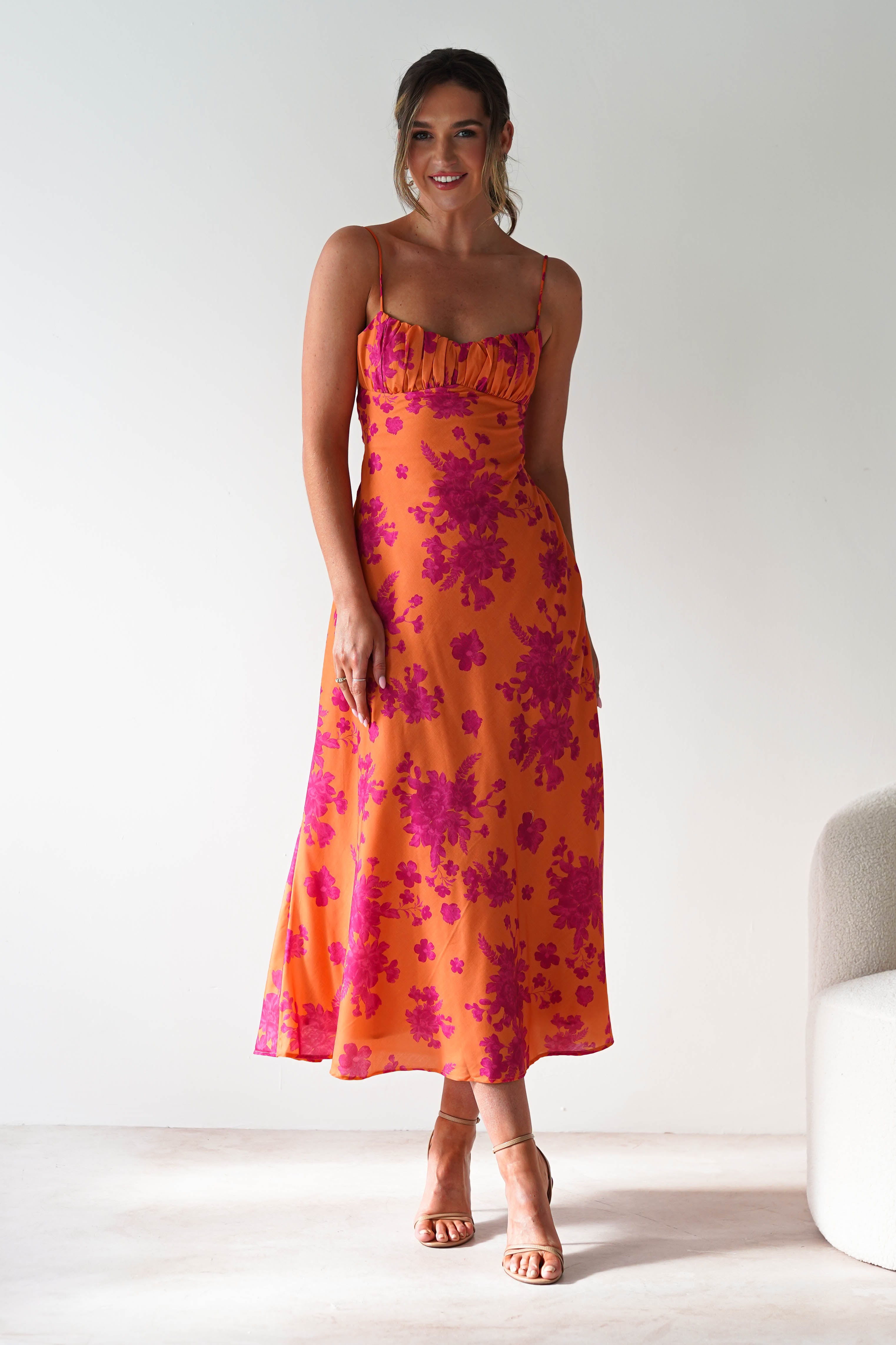 A smiling woman stands indoors in a sleeveless orange wedding guest dress with bright pink floral patterns and strappy heels. She poses with relaxed hands, and there’s a light-colored chair to her right.